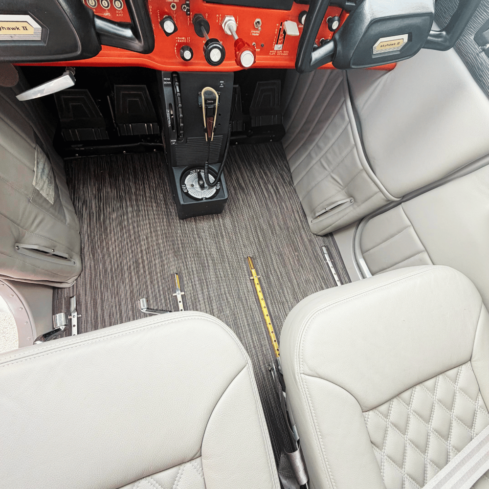 Interior of a small plane with two white leather seats, a red dashboard with various controls, a microphone, and a flooring with a dark striped carpet.