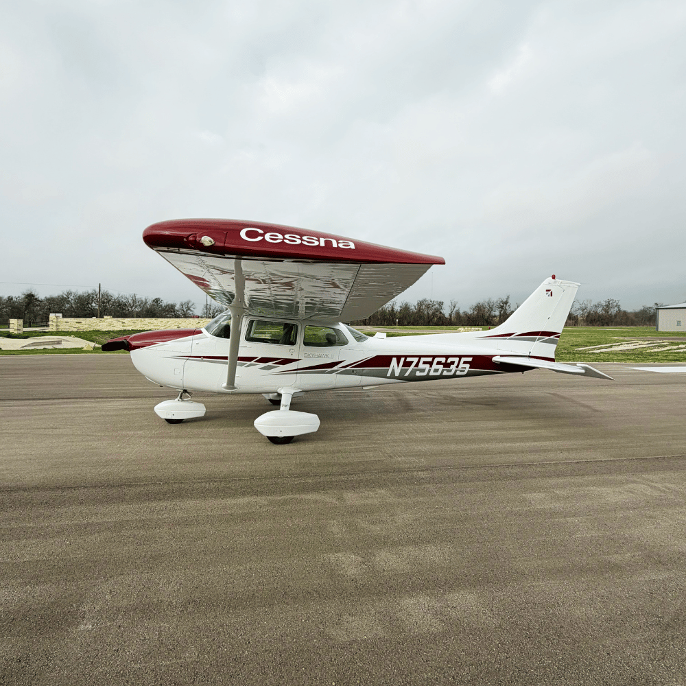 Small Cessna airplane parked on tarmac with red and white exterior, overcast sky in background.