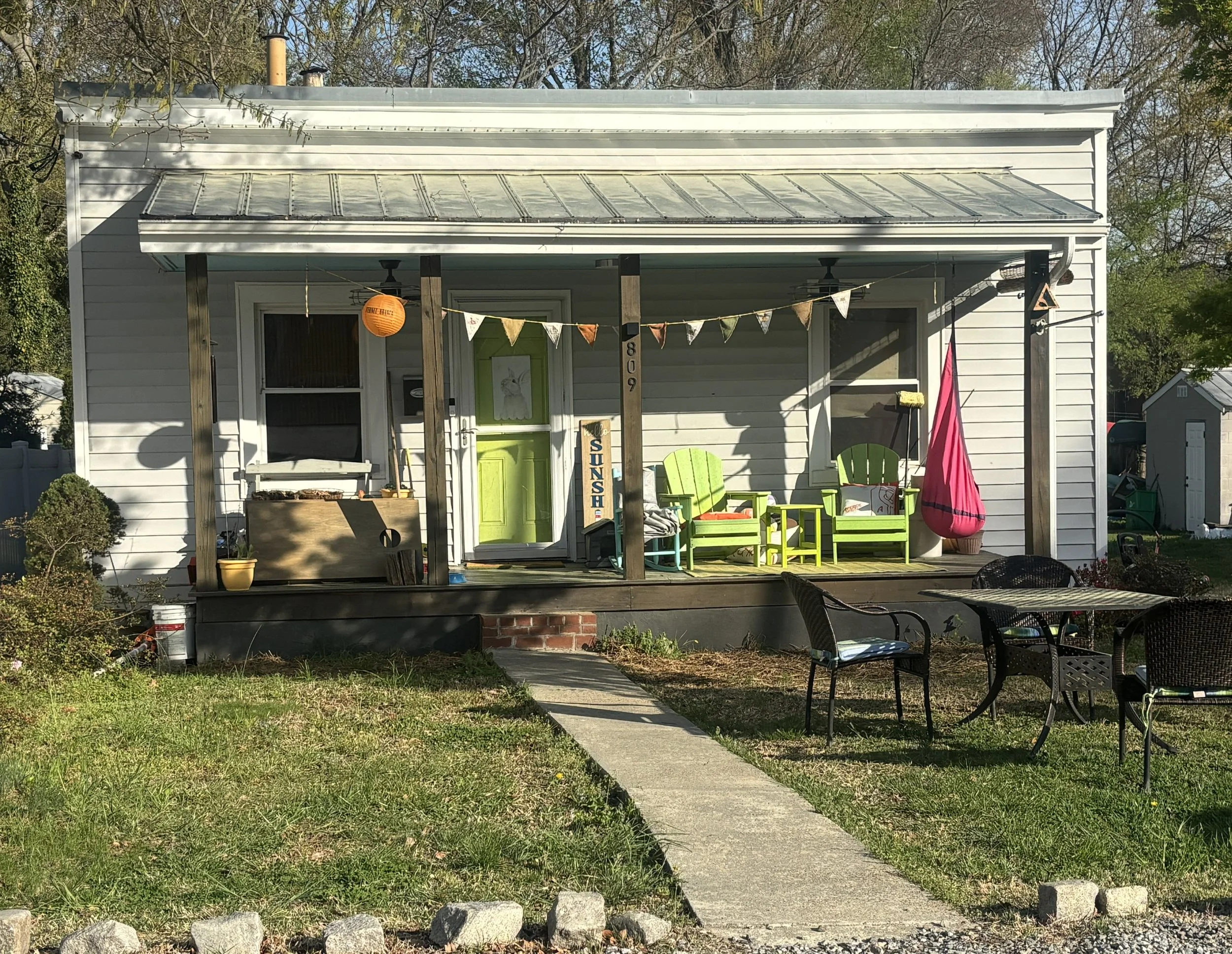Front view of a white house with a small covered porch, plants, colorful chairs, and outdoor decorations, with trees and grass in the background.