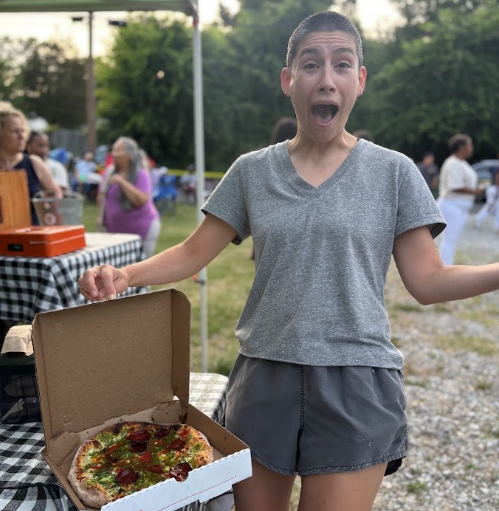 A woman with a surprised expression standing outdoors at a gathering, holding an open pizza box with pizza, surrounded by people and outdoor tables.
