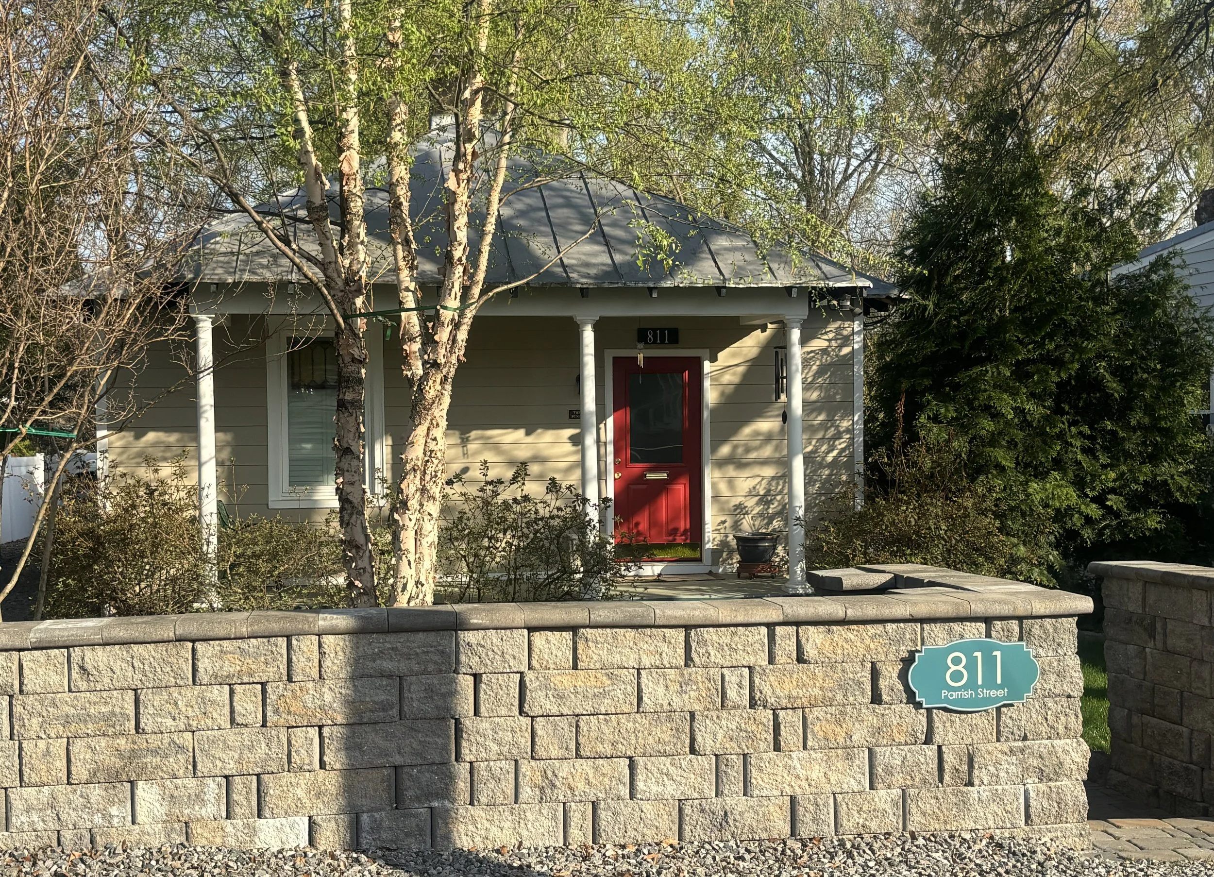 A house with a light yellow exterior, a red door, and a gray roof, surrounded by trees and bushes. There is a stone wall with a blue street sign reading "811 Parrish Street" in the foreground.