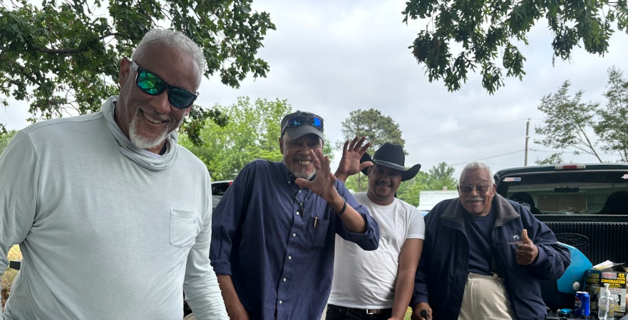 Four men outdoors, smiling, with trees and a truck in the background. One man wears a white long-sleeve shirt and sunglasses, another wears a cowboy hat and white t-shirt, another wears a blue shirt and sunglasses, and the last man wears glasses and a dark jacket.