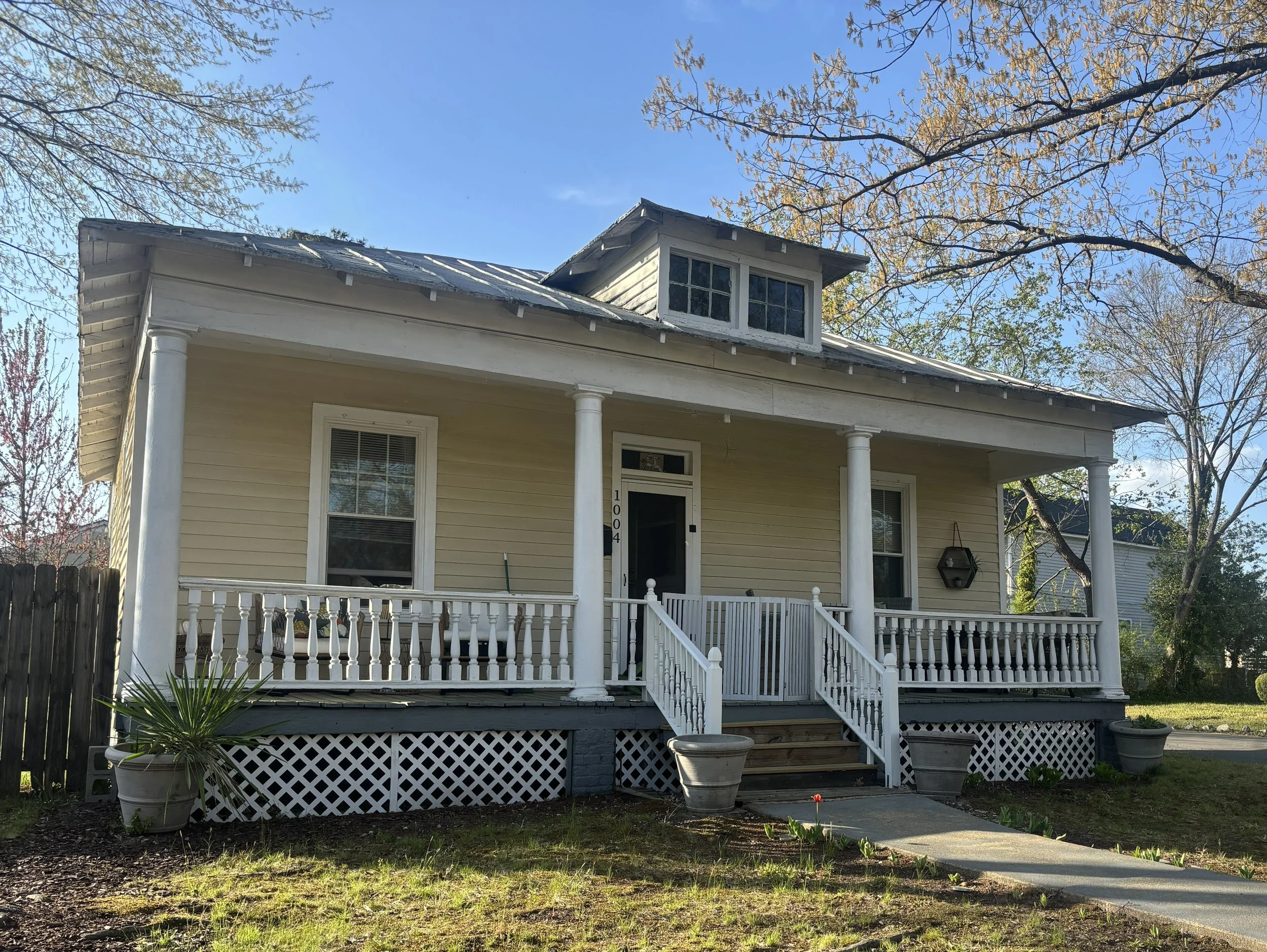 A two-story yellow house with white trim and a front porch with white railing. The house has large windows and a small dormer window on the roof. There are trees around the house with some leaves turning color, indicating fall. Two large planters with plants are on the front porch, and a small path leads to the stairs.