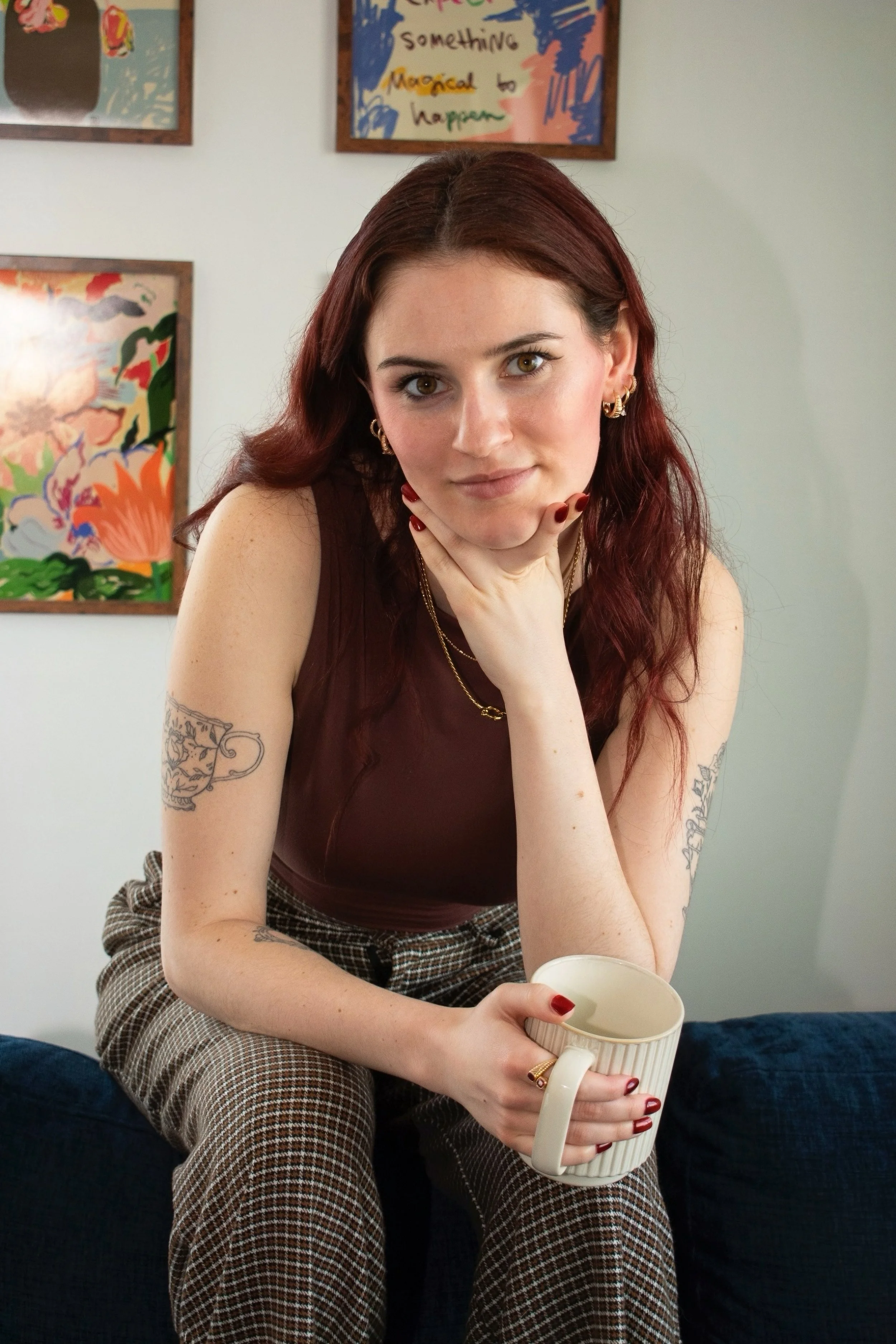 A young woman with red hair and tattoos on her arms sitting on a dark couch, holding a white mug with a textured surface. She is wearing a sleeveless dark brown top and patterned pants, with jewelry including earrings, rings, and necklaces. She rests her chin on her left hand and looks directly at the camera. The background includes colorful framed artwork on a white wall.