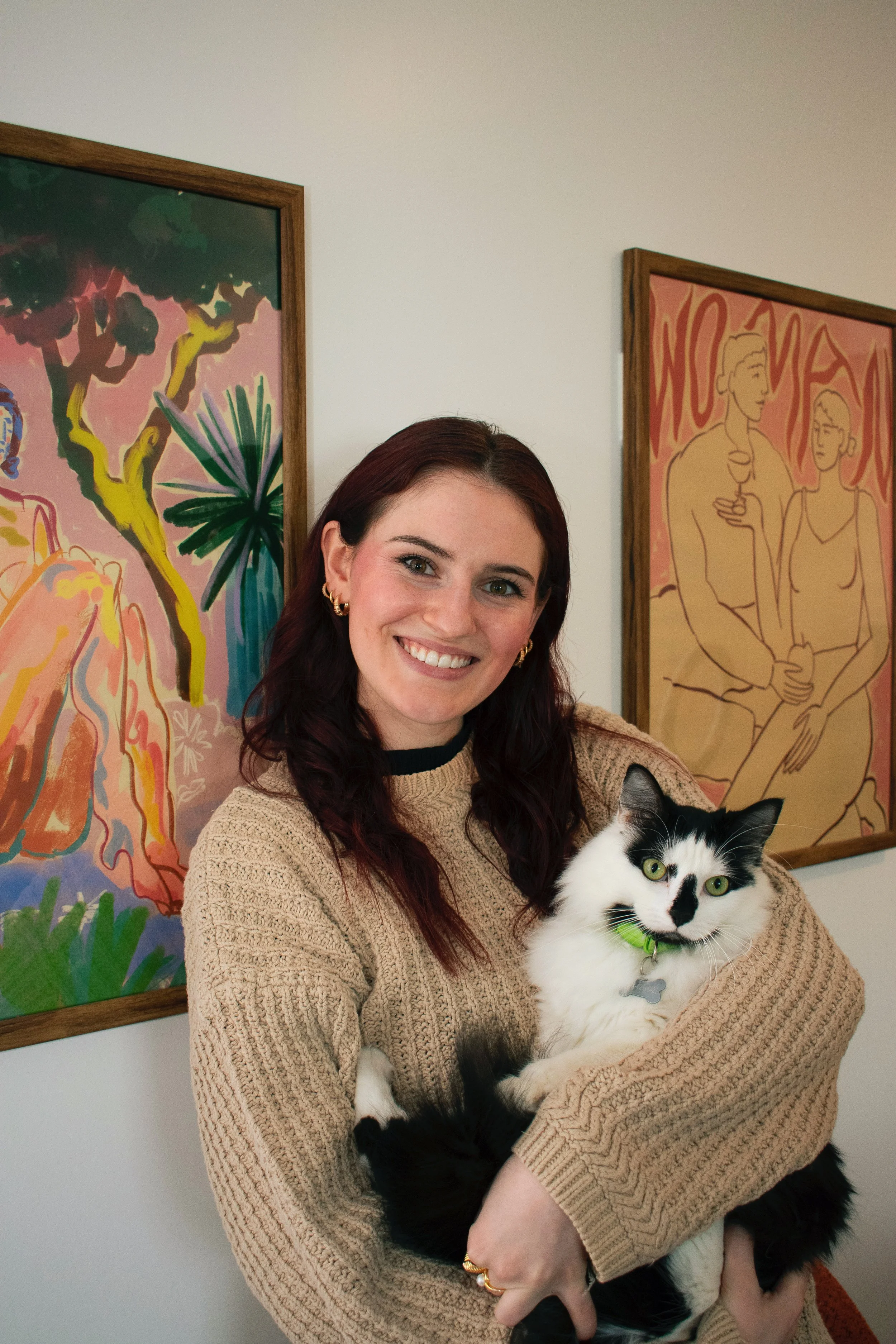 A woman with dark red hair smiling and holding a black and white cat in her arms, standing in front of colorful artwork on a wall.