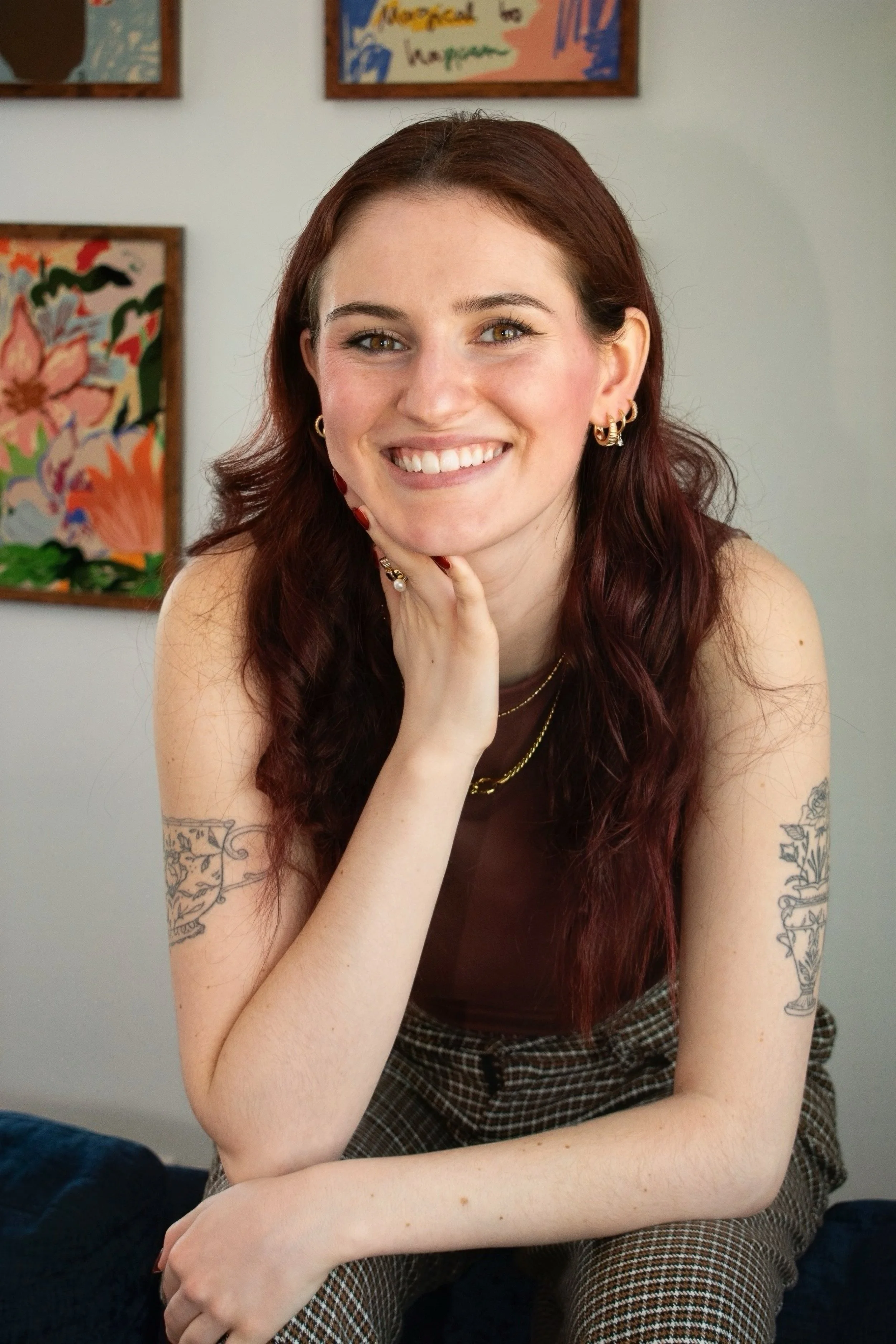 A young woman with long wavy red hair, smiling, sitting indoors with her hand resting on her chin. She has tattoos on her arms and is wearing gold jewelry, earrings, and a sleeveless top. Behind her, there are colorful artwork and signs on the wall.