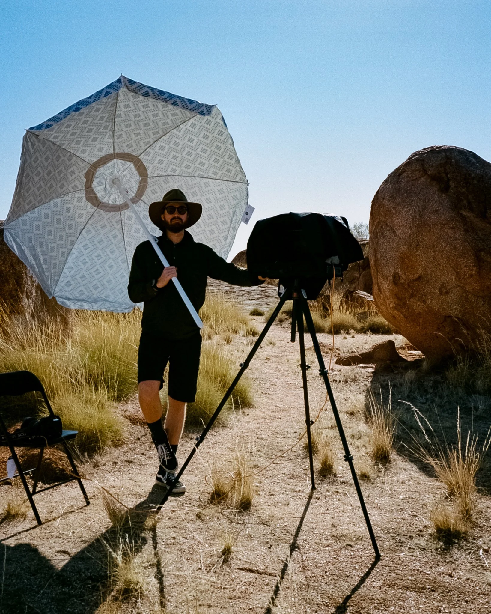 A man standing outdoors in a desert landscape with rocks and dry grass, holding a large patterned umbrella and standing next to a camera on a tripod, wearing sunglasses, a wide-brimmed hat, a black shirt, shorts, and sneakers.