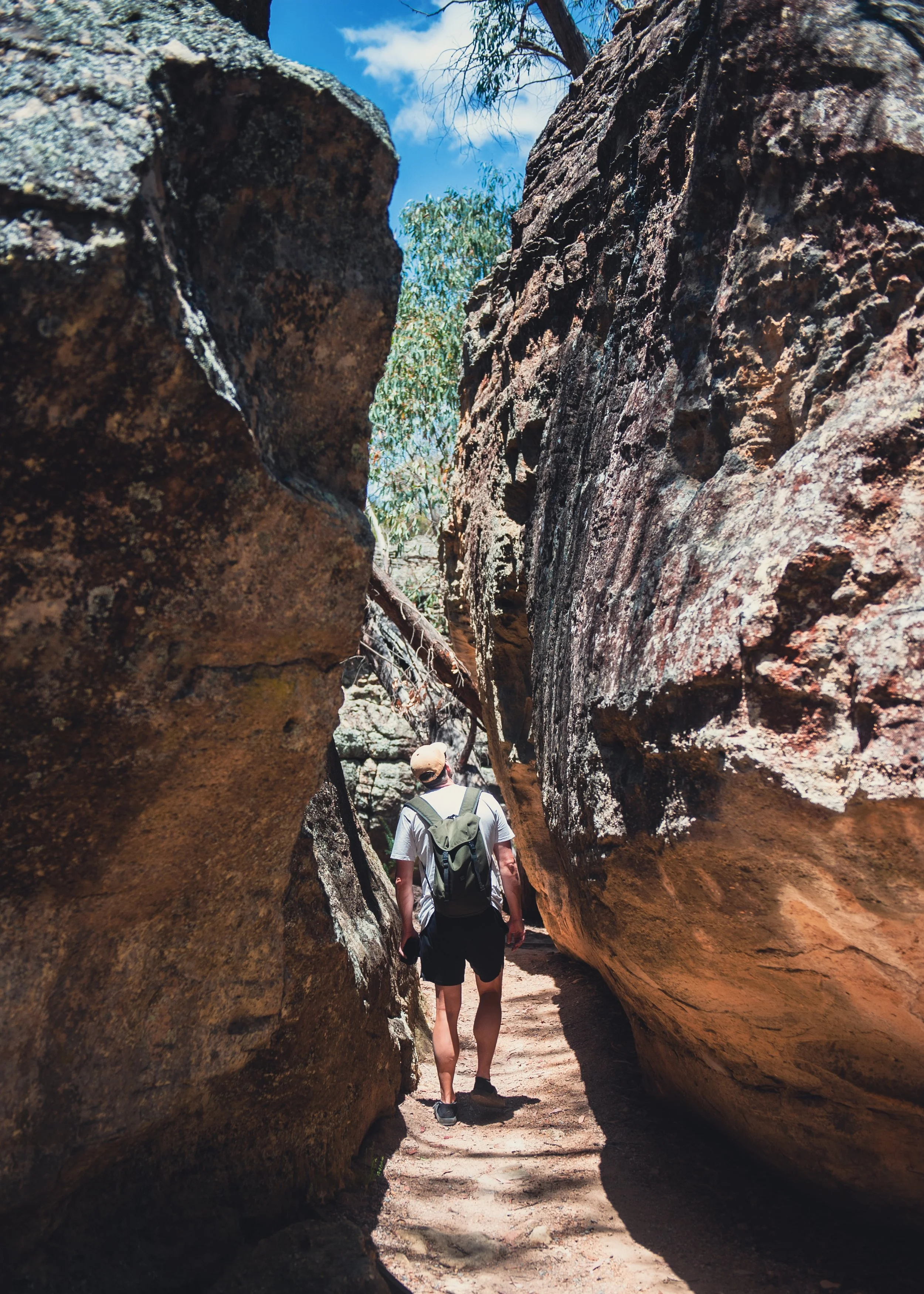 A hiker walking between large rock formations on a trail in a desert landscape under a blue sky.