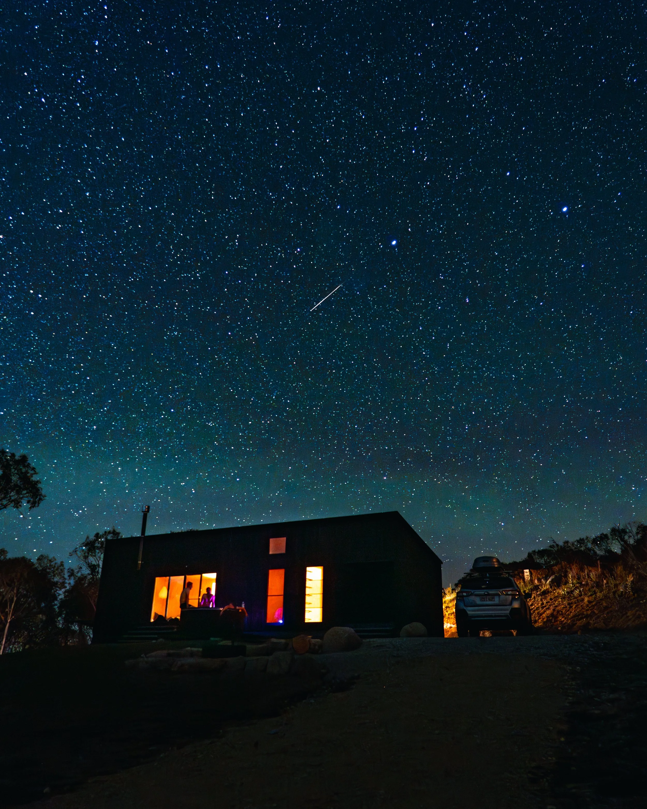 A house at night with illuminated windows, a car parked outside, and stars in the sky including a shooting star.