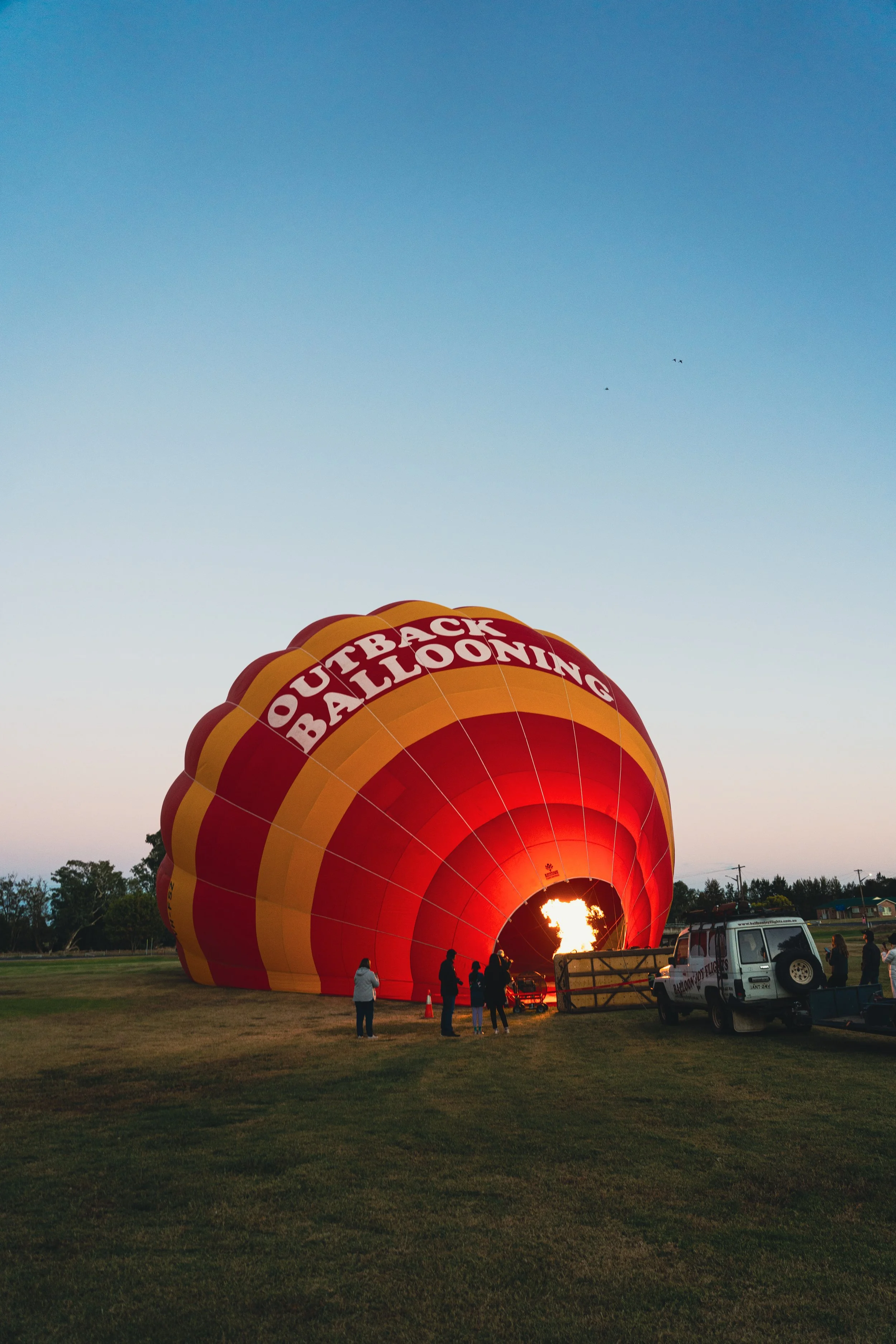 A hot air balloon labeled 'Outback Ballooning' is being inflated on a grassy field at dawn or dusk, with a flame coming from the burner inside the balloon and a few people standing nearby.