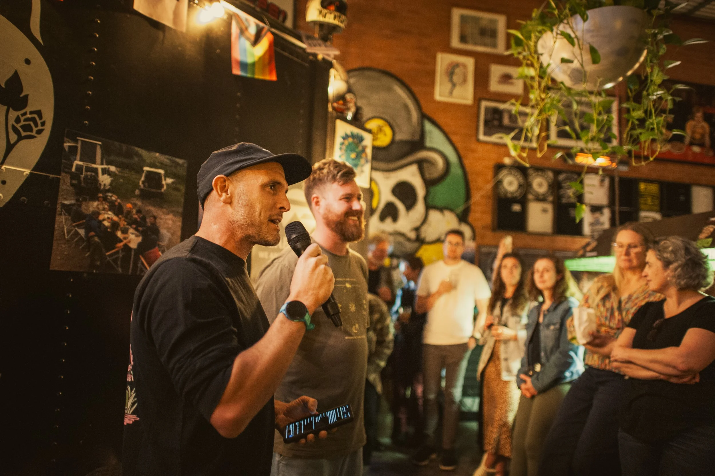 A man in a black cap and black shirt speaking into a microphone at a gathering, while another man beside him listens. Several people stand in the background, some holding drinks. The setting is a dimly lit room with decorations including framed photos, a large mural of a skull with a green bandana, and a small rainbow flag.
