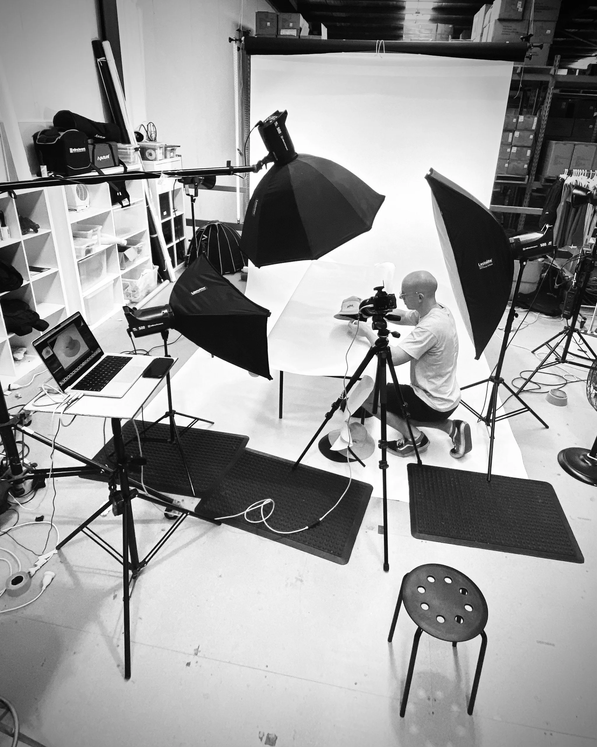 A photographer setting up a photo shoot in a studio with a white background, professional lighting, and a laptop, in a space with shelves filled with boxes and equipment.