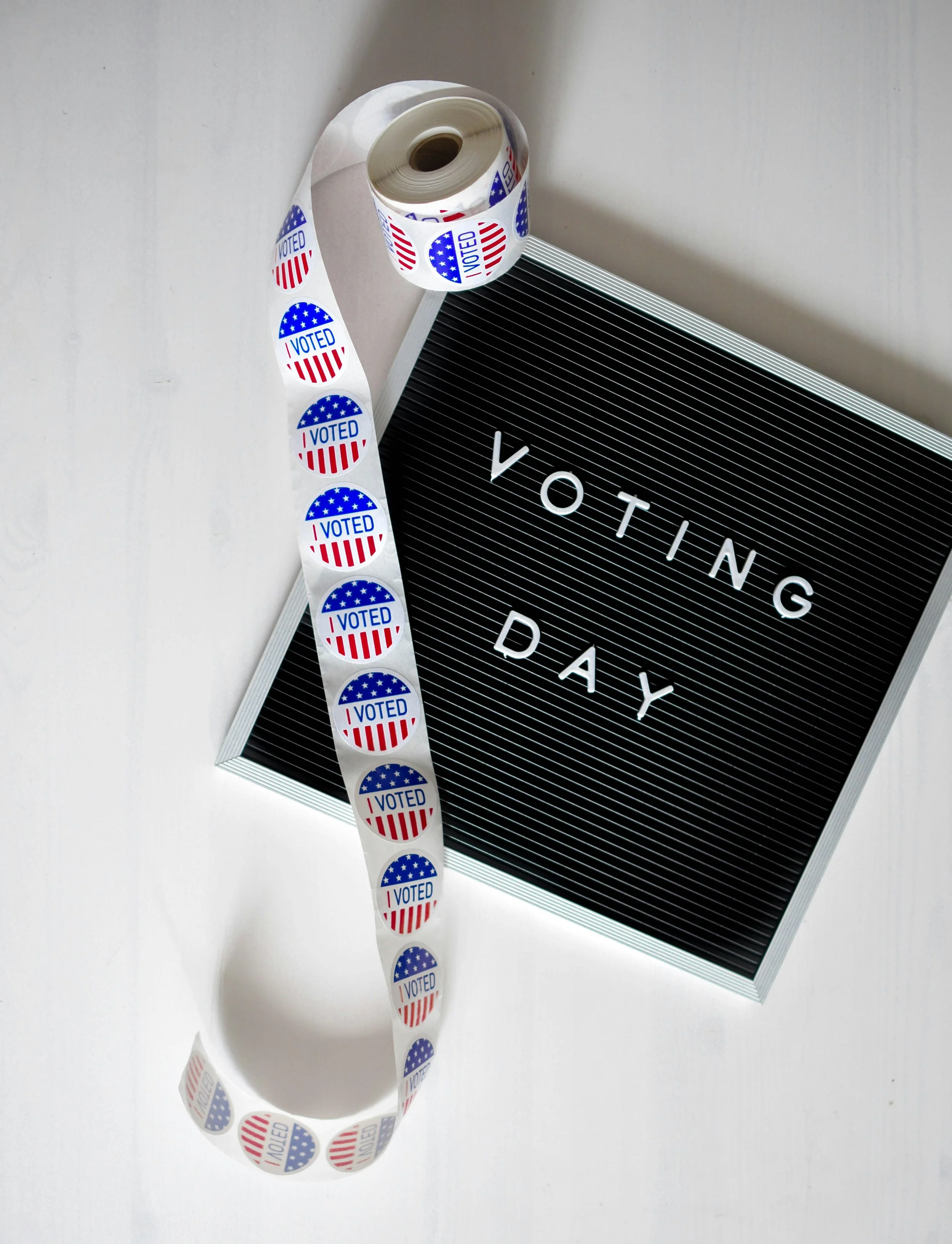 A roll of sticker labels with a patriotic American theme that say "I VOTED" and have red and white stripes and blue with white stars, lying beside a black letter board with the words "VOTING DAY" in white letters.