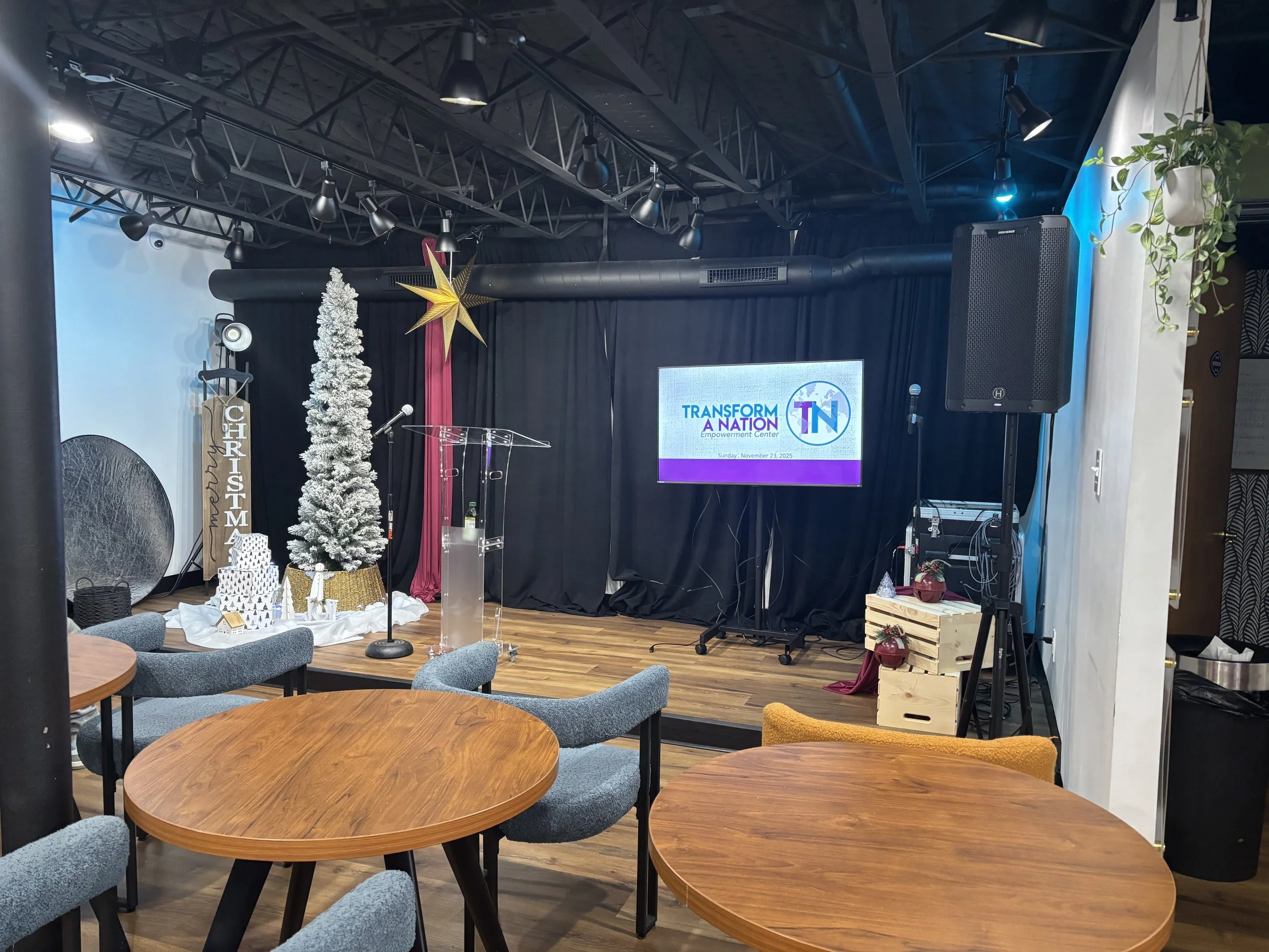Event stage decorated with Christmas themed decorations, a white Christmas tree, a golden reindeer, a large star, and a sign that says 'CHRISTMAS'. There is a large screen on the right side displaying the logo 'Transform a Nation'. Seating area with round wooden tables and gray chairs in the foreground.