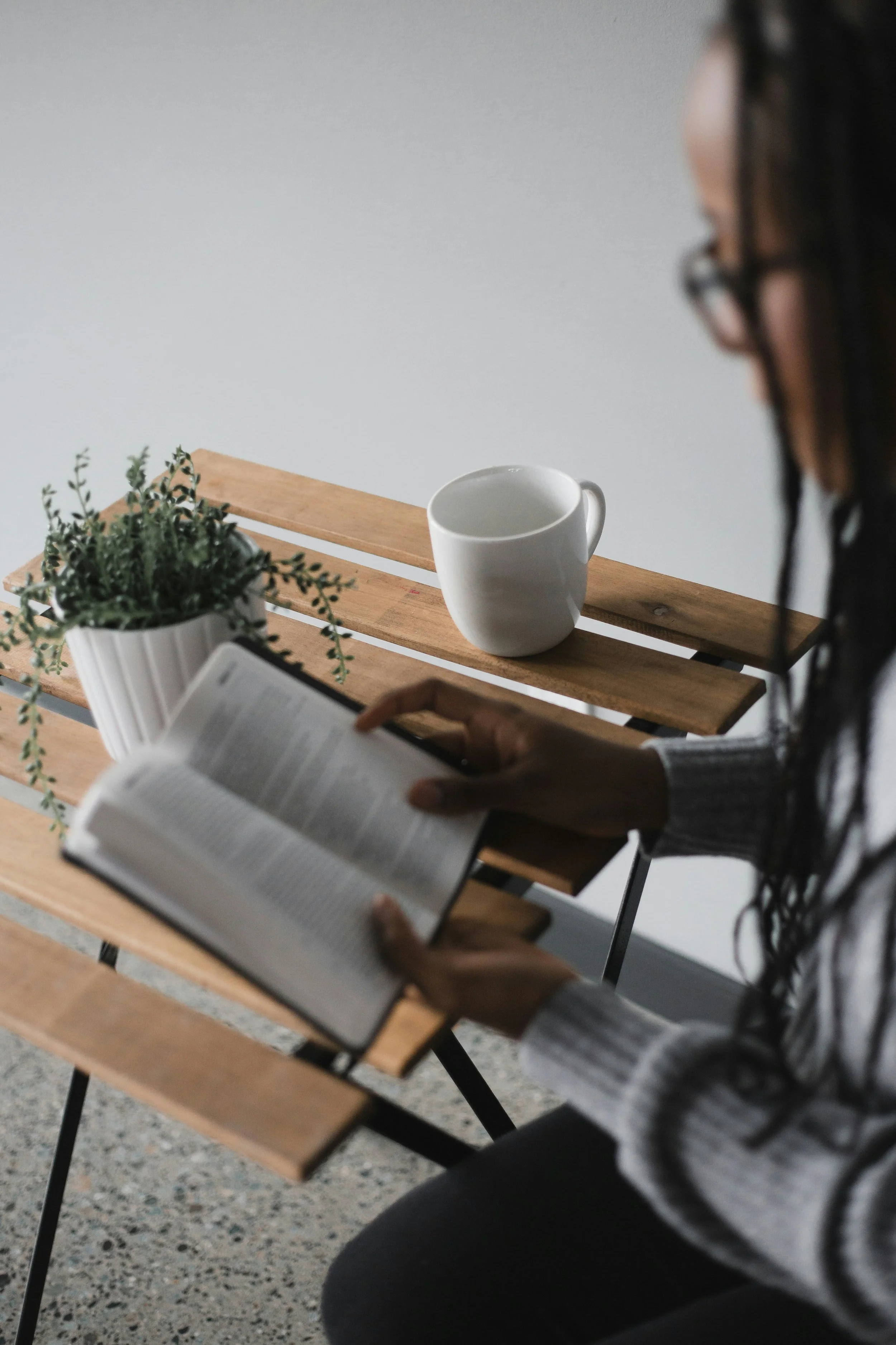 A person with long dark hair and glasses sitting at a wooden table reading a book, with a white mug and a potted plant on the table.