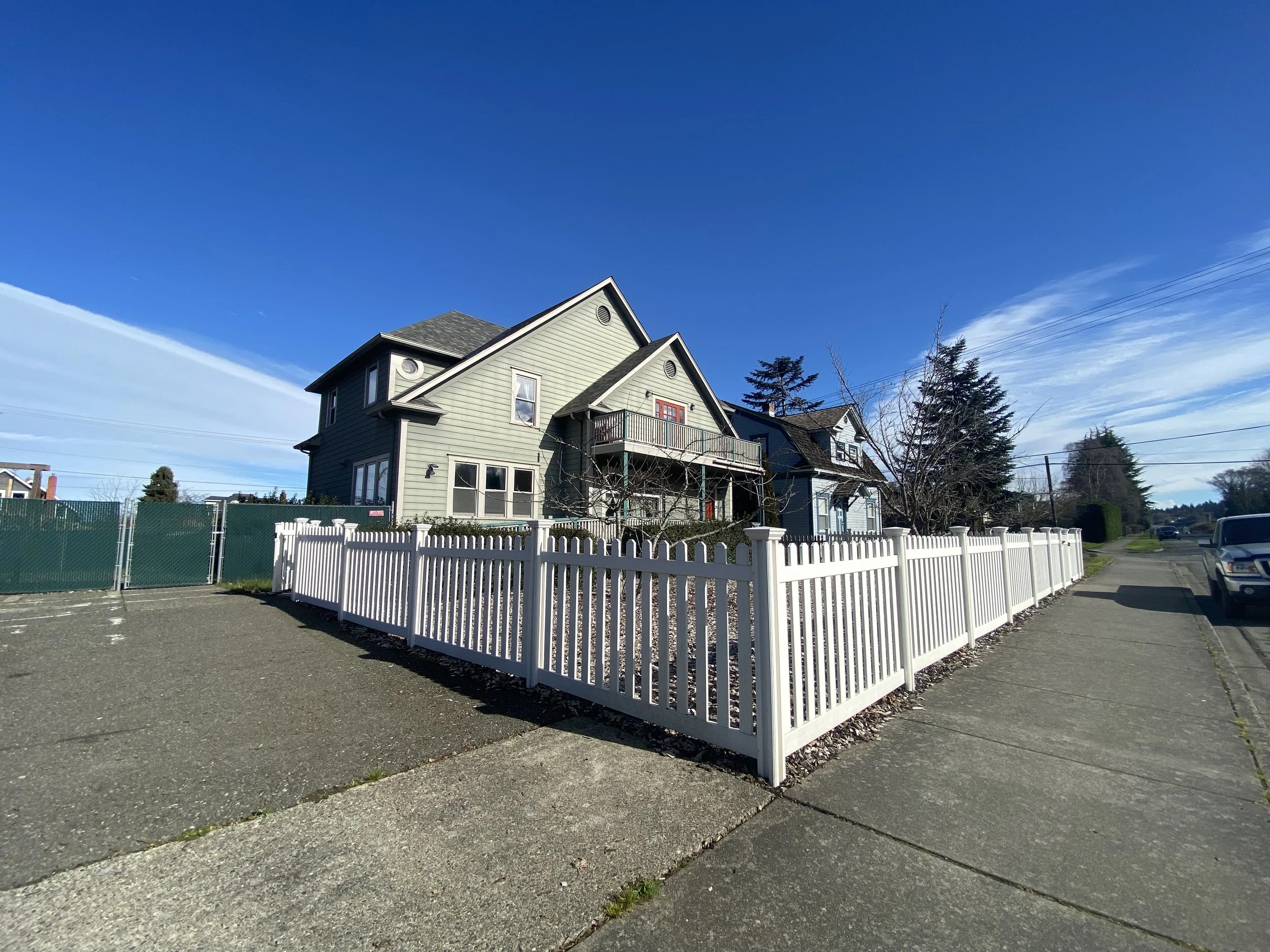 A residential house with beige siding and a small balcony, surrounded by a white picket fence, set against a bright blue sky with a few clouds and trees in the background.