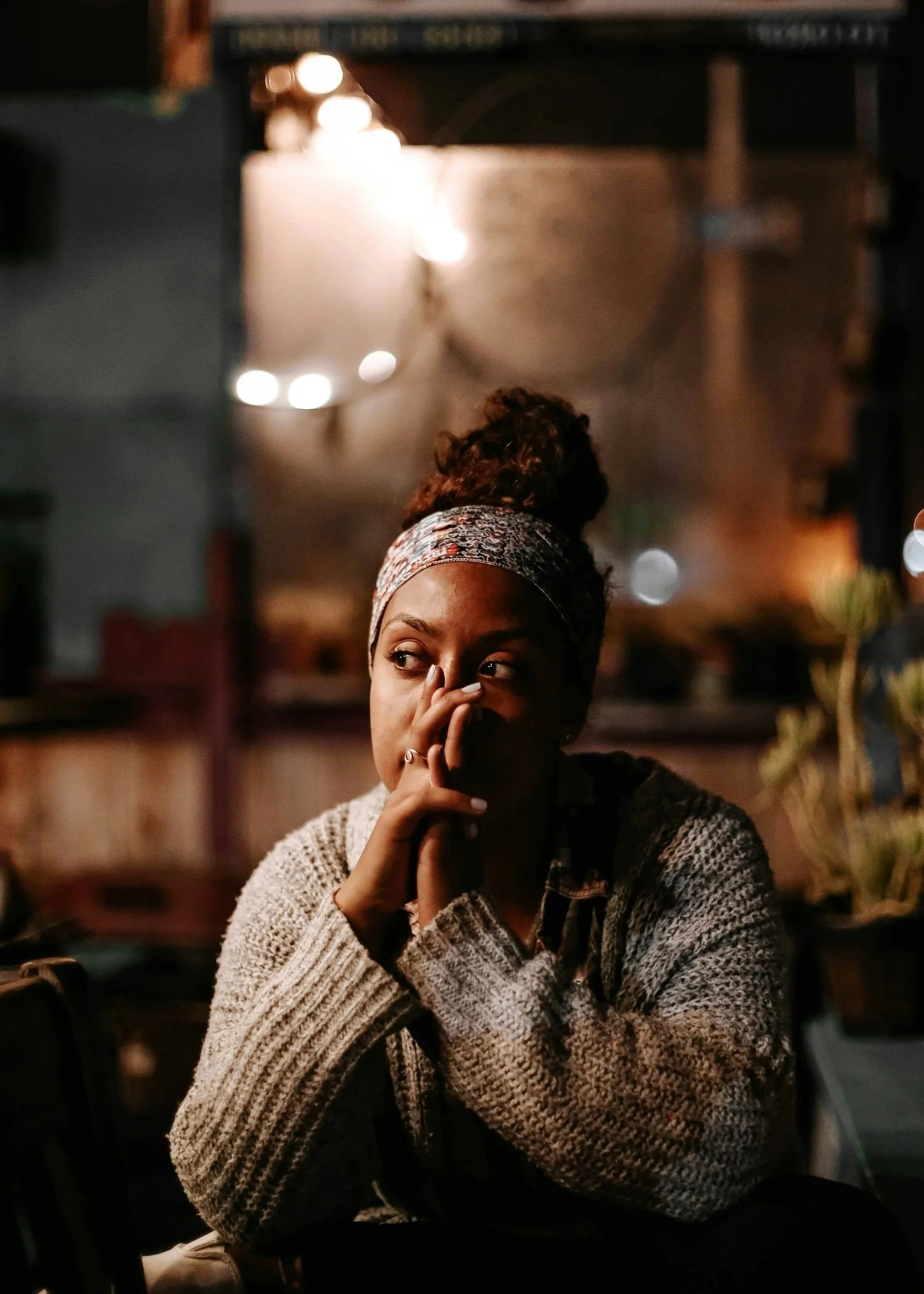 A woman with curly hair wearing a patterned headband and a knitted sweater, sitting indoors at night with her hand covering her mouth, and looking to her right.