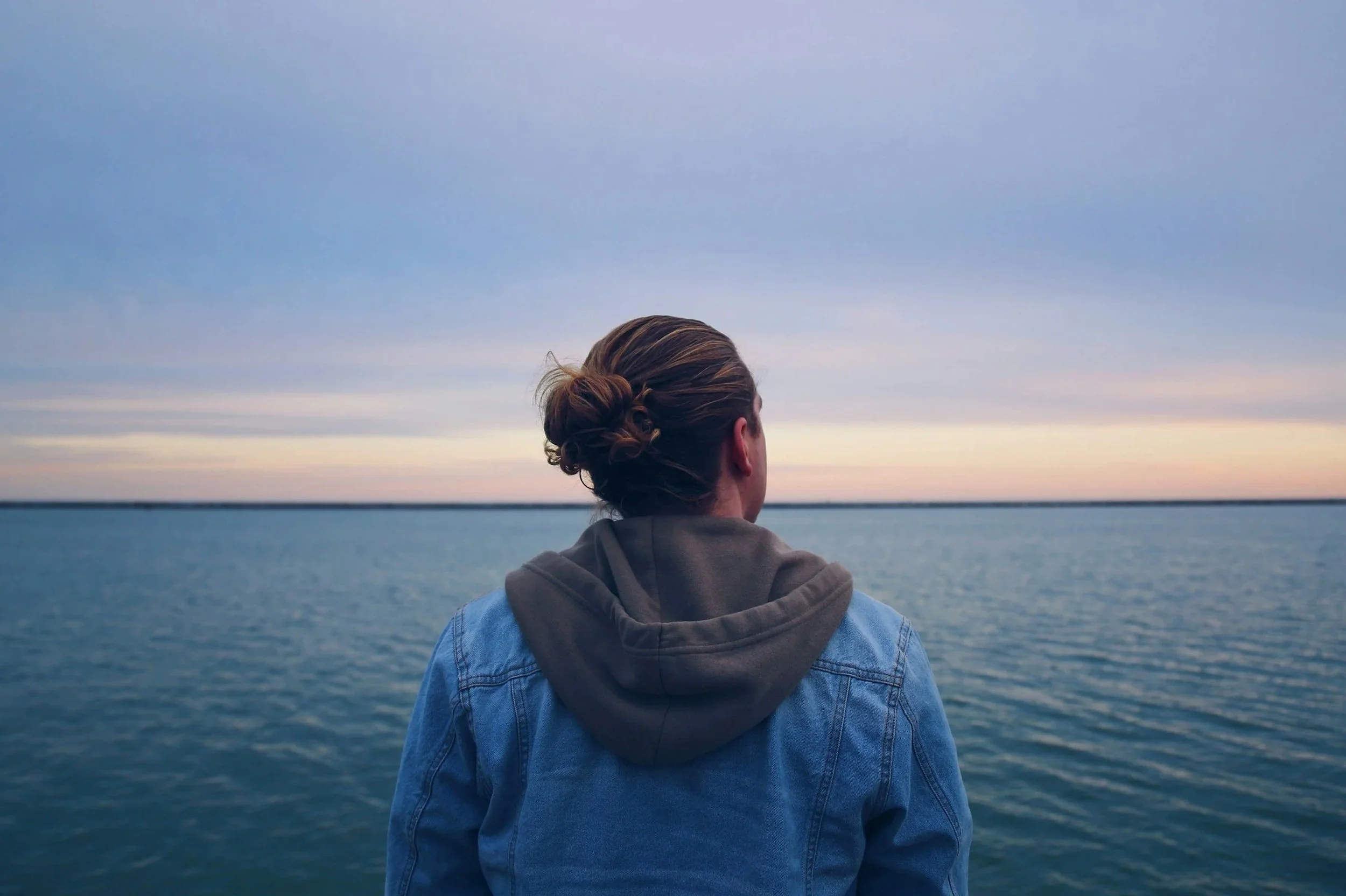 Person with brown hair tied in a bun wearing a denim jacket and gray hoodie, looking out at a large body of water with an expansive sky at sunset or sunrise.