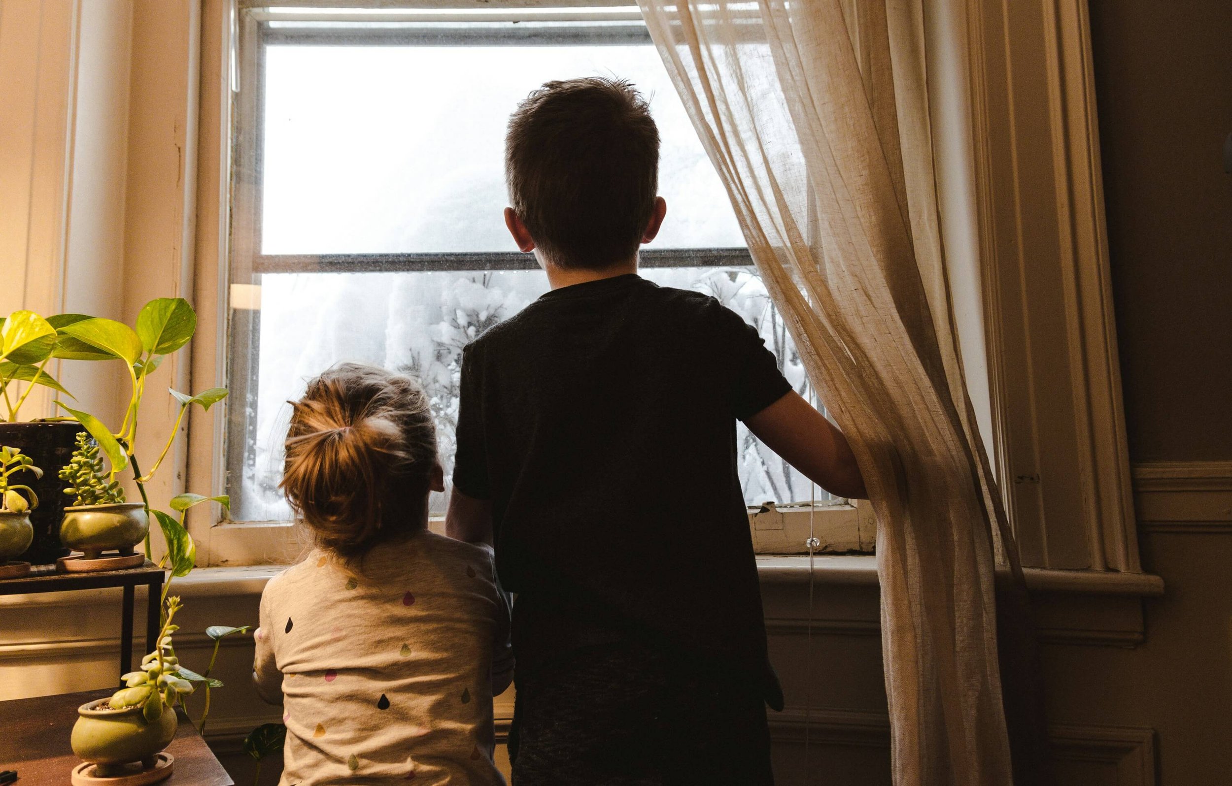 Three children, two girls and a boy, look out a window during a snowy day inside a cozy room. The girl in front has her hair in a bun, the boy stands with his back to the camera, and the girl beside him has short hair. Potted plants are on the windowsill, and a beige curtain partially covers the window.
