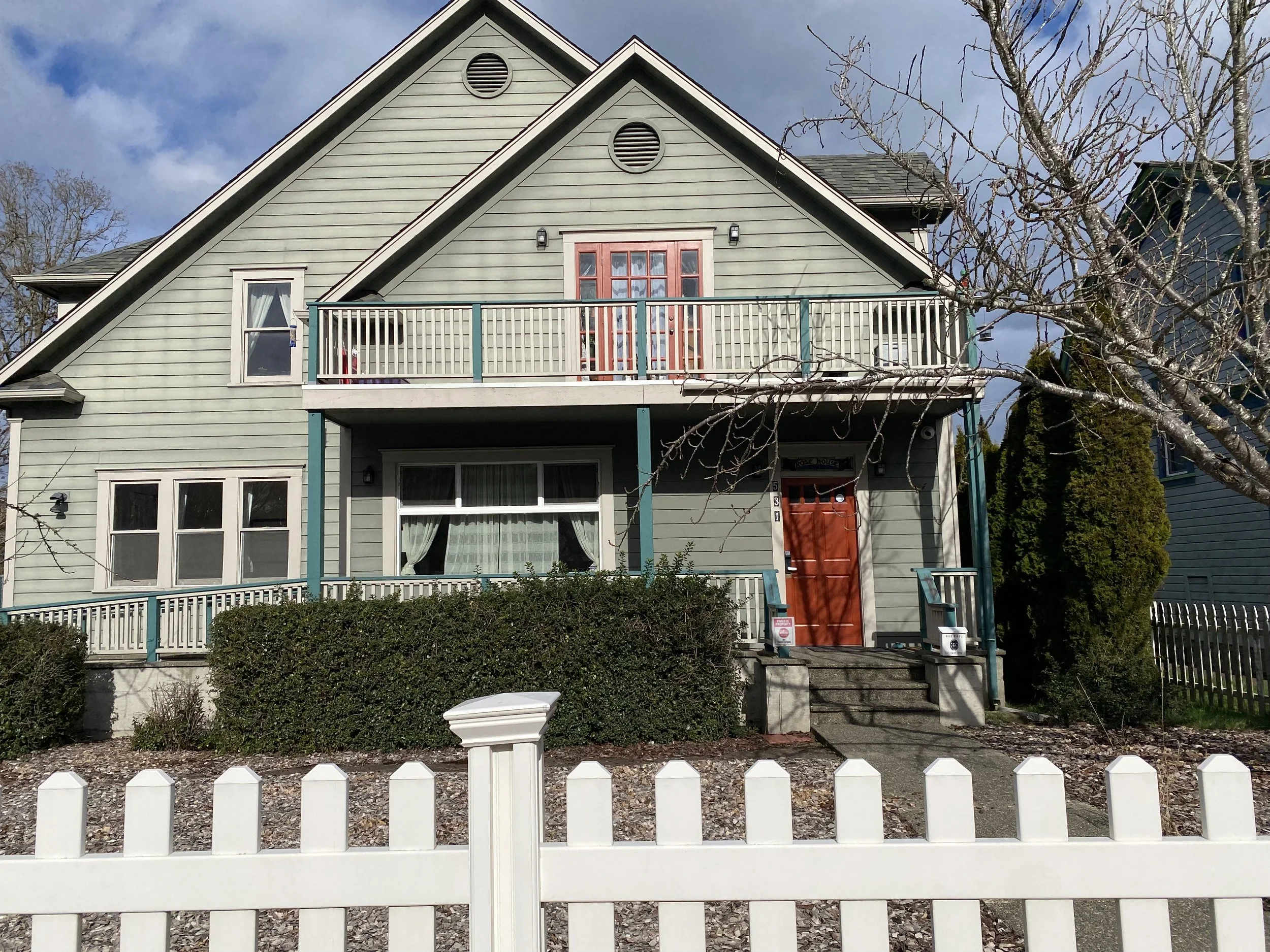 Front view of a two-story house with a white picket fence, green shingles, a small balcony with a door, and leafless trees in the front yard. Overcast sky in the background.