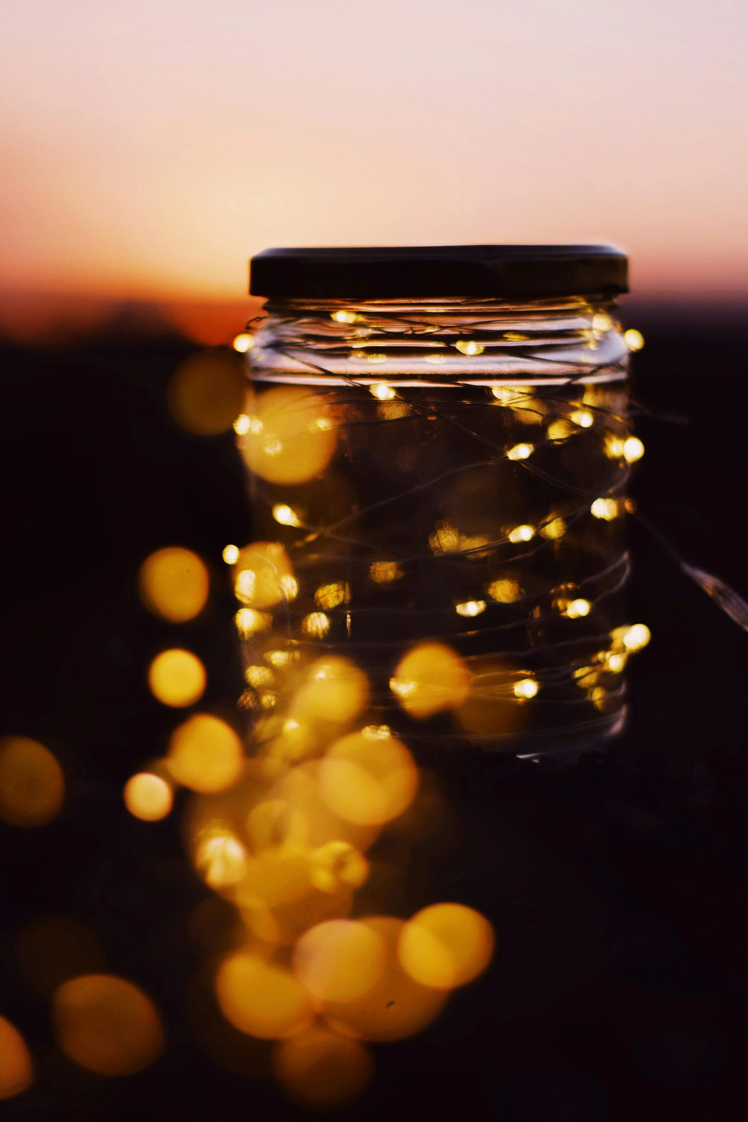 Close-up of a glass jar with a black lid, decorated with a string of tiny, warm-colored fairy lights, reflecting on a dark surface with a blurred sunset or sunrise in the background.