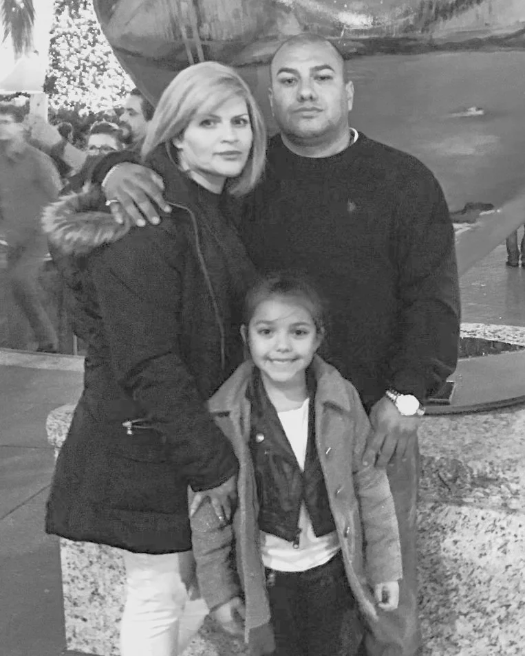 A group of three people, a woman, a man, and a young girl, standing close together outdoors at night, smiling at the camera. The woman has shoulder-length hair and is wearing a coat. The man is wearing a dark sweater and a watch. The girl has long hair and is dressed in a jacket. Christmas lights and trees are visible in the background.