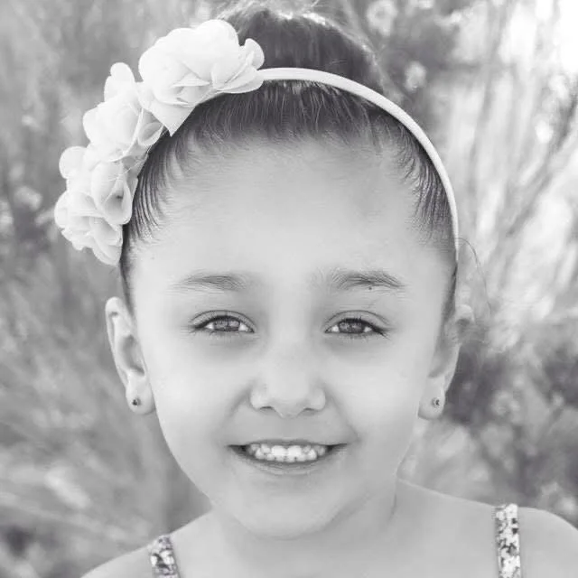 Black and white photo of a young girl smiling, wearing a headband with flowers and earrings, outdoors with blurred palm trees in the background.