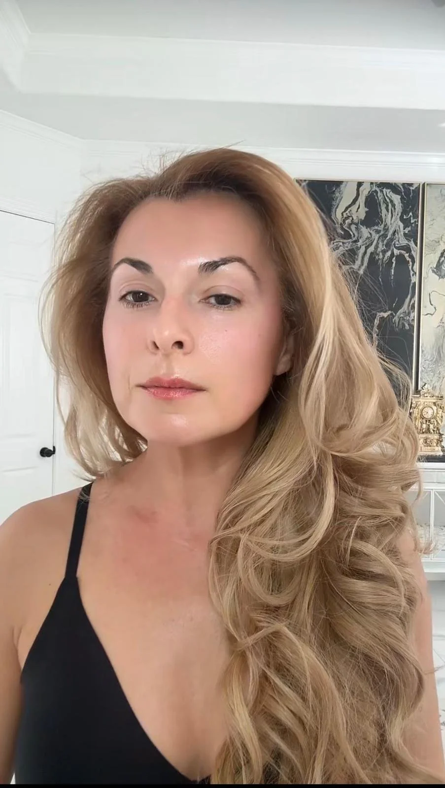 Close-up of a woman with long, wavy blonde hair, wearing a black top, standing in a room with artwork on the wall.