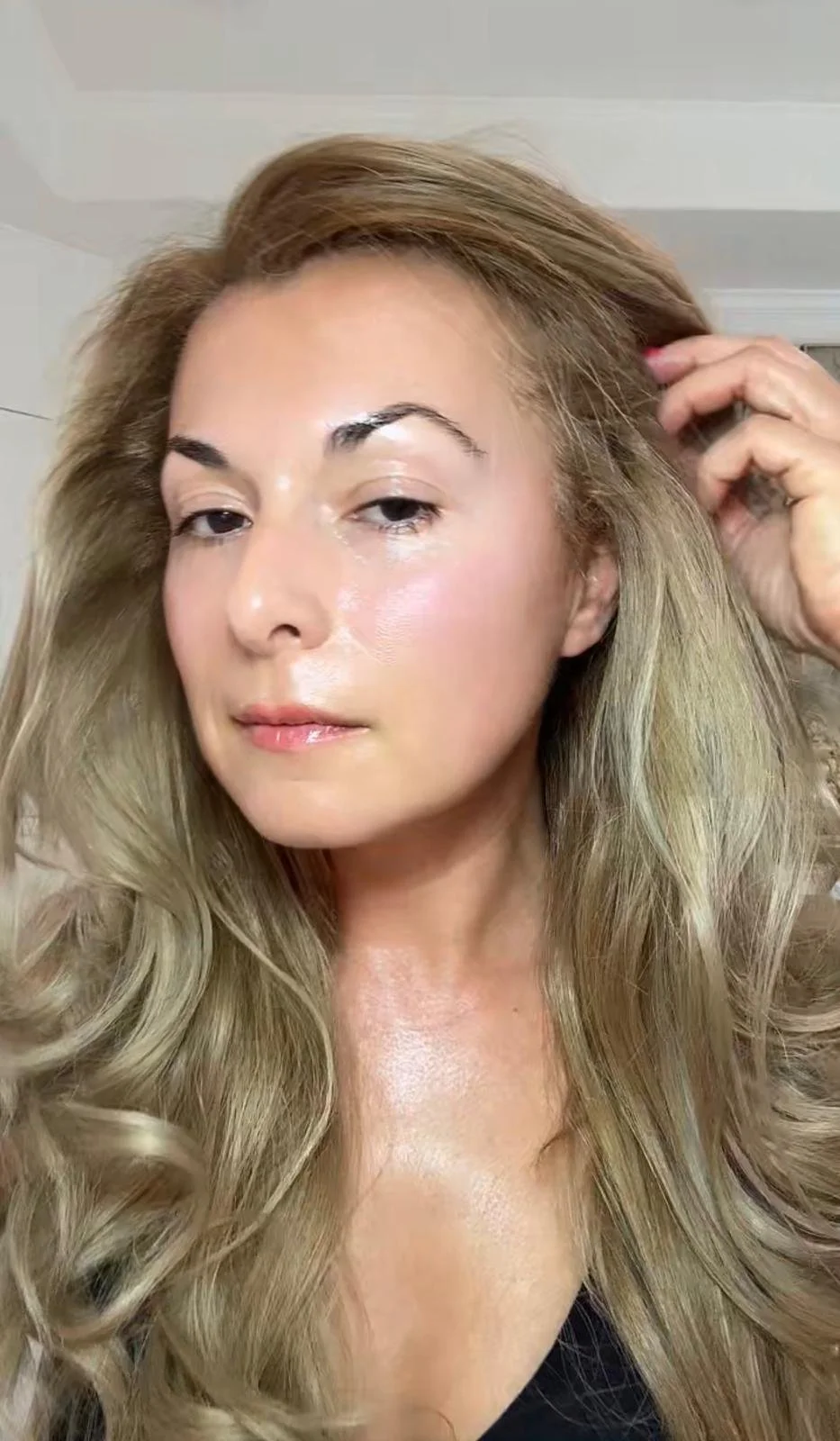 Close-up of a woman with long, wavy blonde hair, fair skin, and makeup, touching her hair with her right hand.