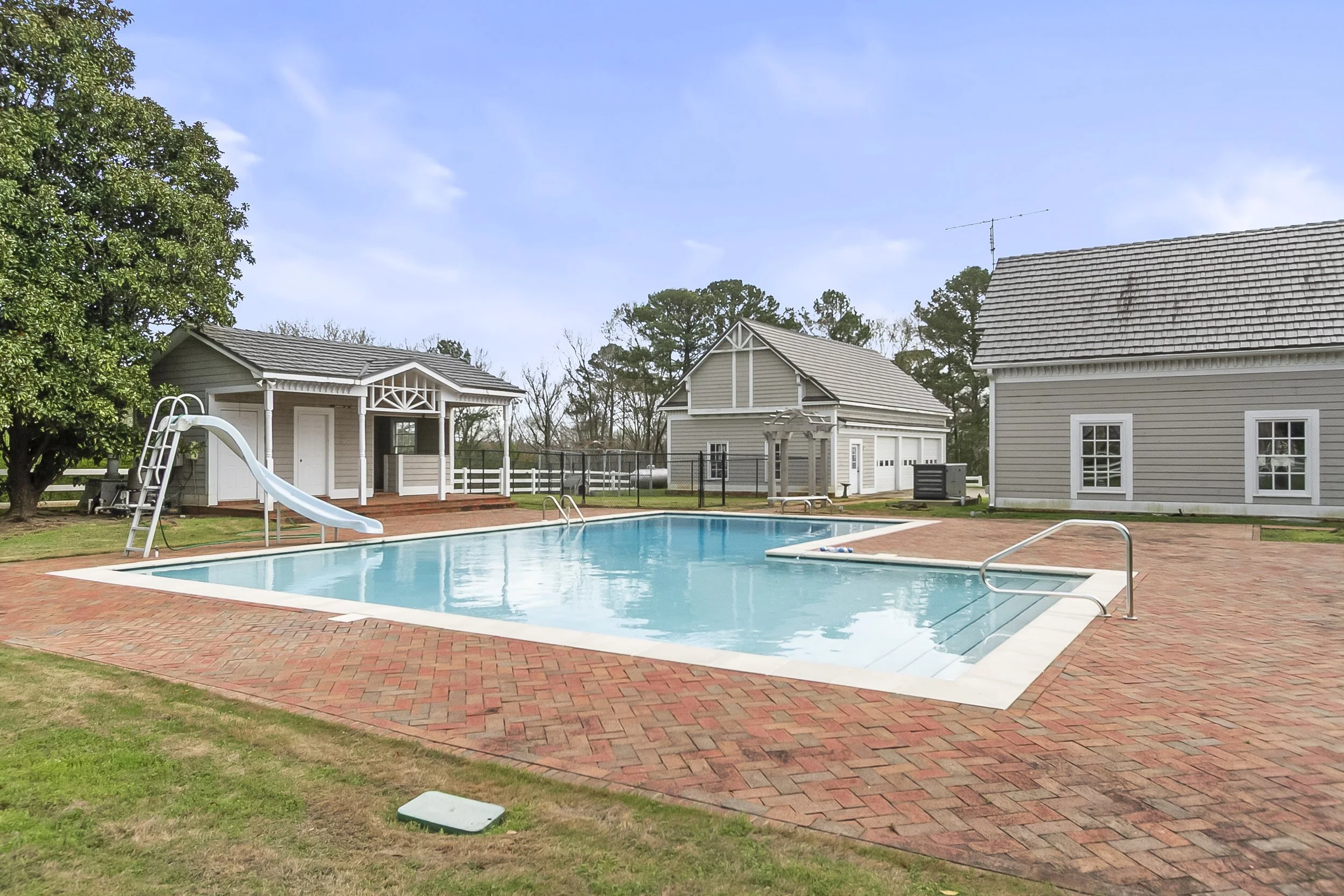 A backyard with a swimming pool, a red brick patio, and three small buildings or sheds. There is a slide and a ladder on the left side of the pool, and a small covered structure in the center backyard. The sky is partly cloudy.