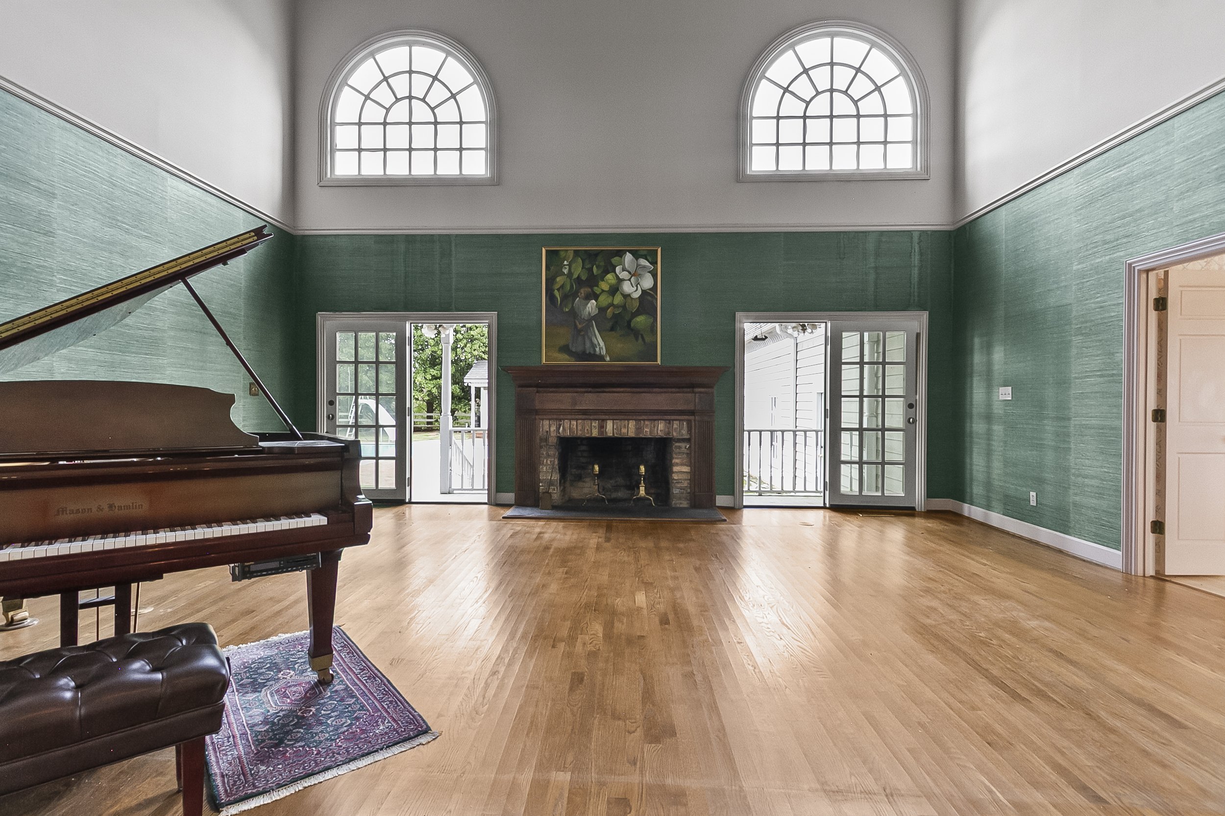 Spacious living room with green walls, hardwood floors, a brick fireplace, large windows, and French doors leading outside. Piano and stool are in the left corner, with artwork above the fireplace.