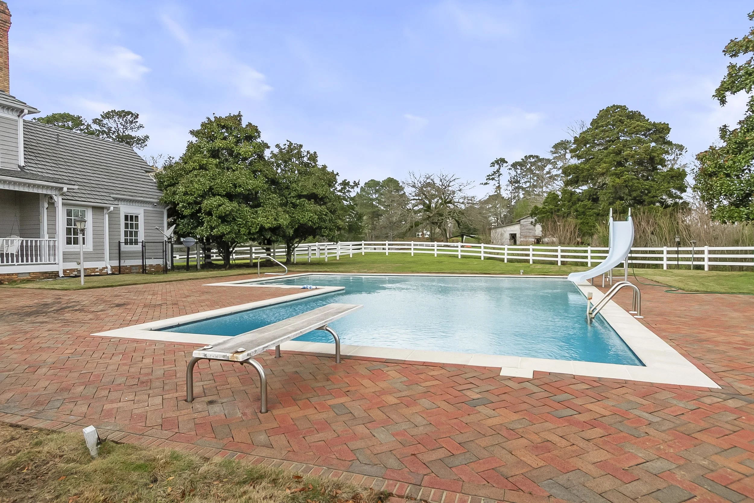 An outdoor swimming pool with a water slide on the right side, surrounded by a brick patio and green grass. There are trees and a white fence in the background, and part of a house on the left.