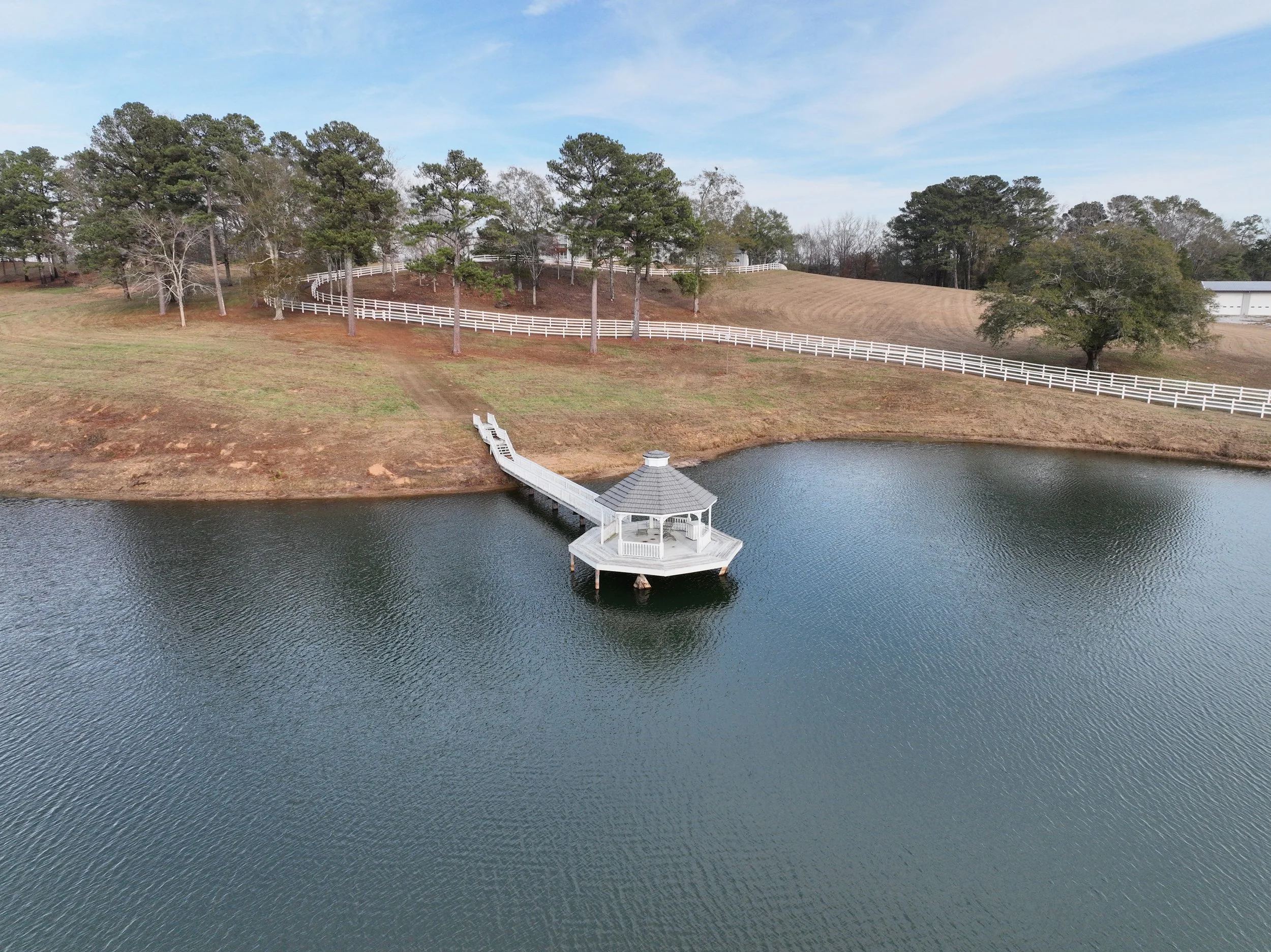 A small gazebo at the end of a dock extending into a lake, with a hillside and trees in the background, under a partly cloudy sky.