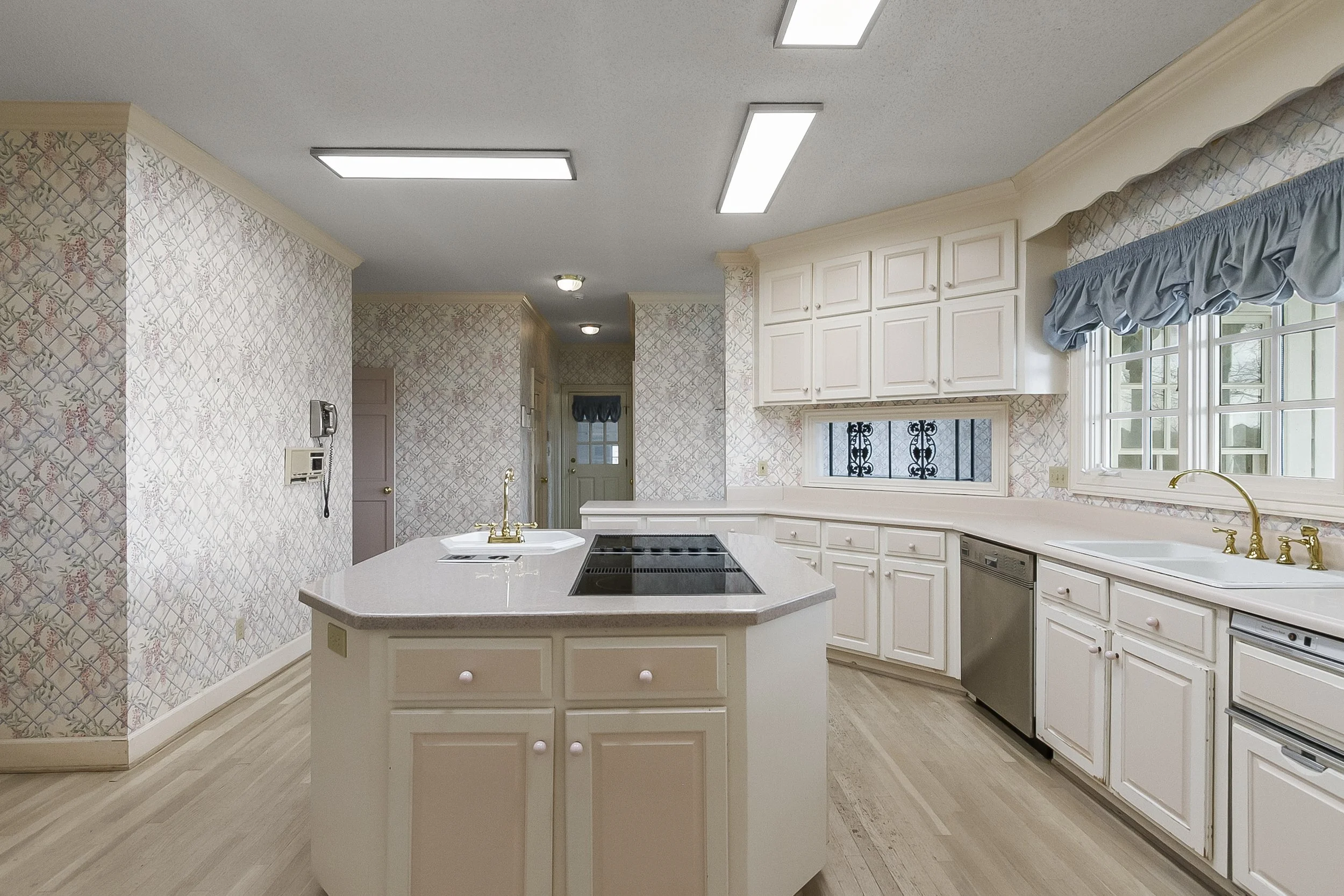 A kitchen with white cabinets, a white island with a built-in stovetop and sink, light wood flooring, and a window with a blue valance.