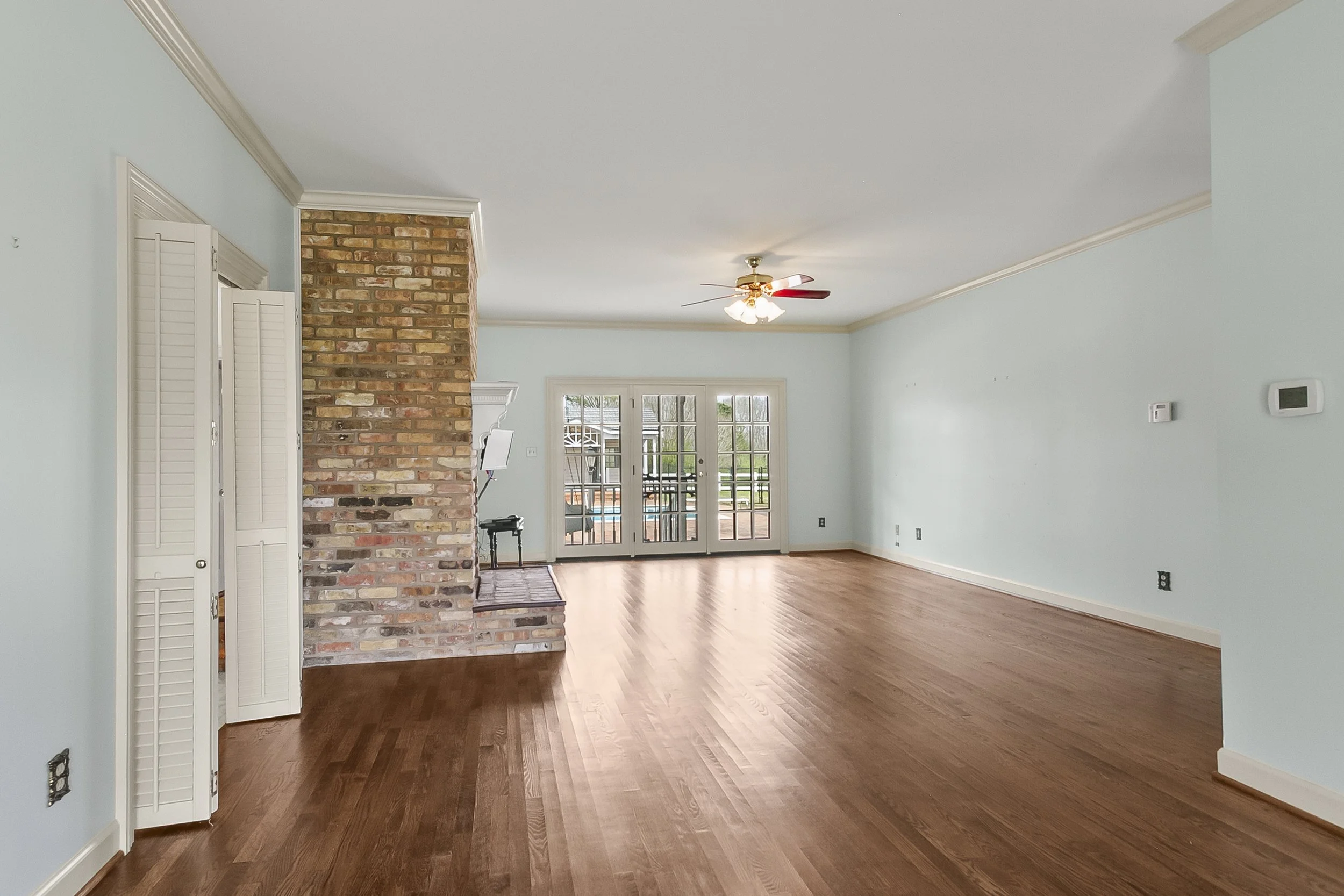 Empty living room with hardwood floors, light blue walls, a ceiling fan, and French doors leading outside. There's a brick chimney and a small black piano near the chimney.