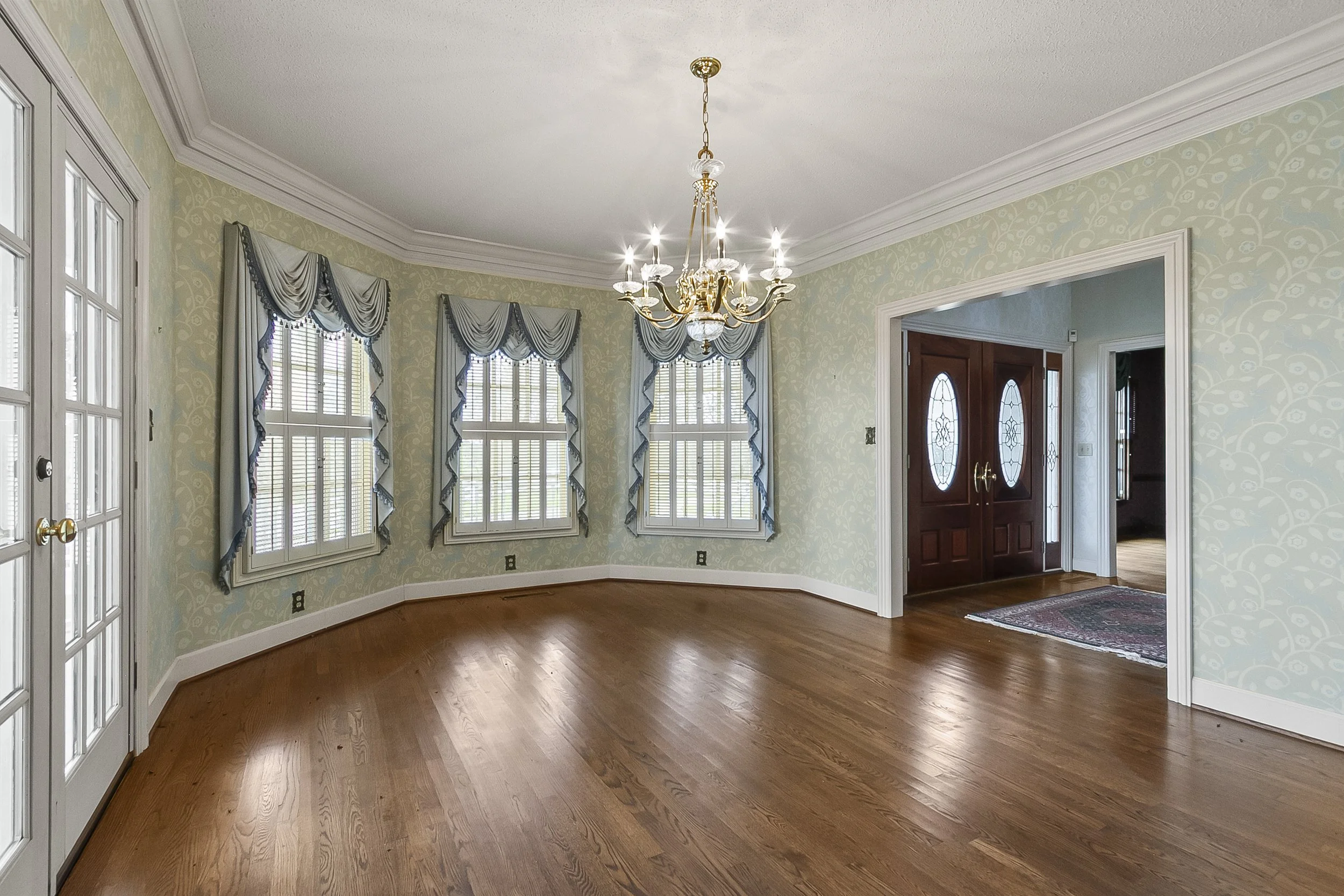 Empty dining room with three large windows with draped valances, hardwood floors, chandelier, and double front doors with oval glass panes.