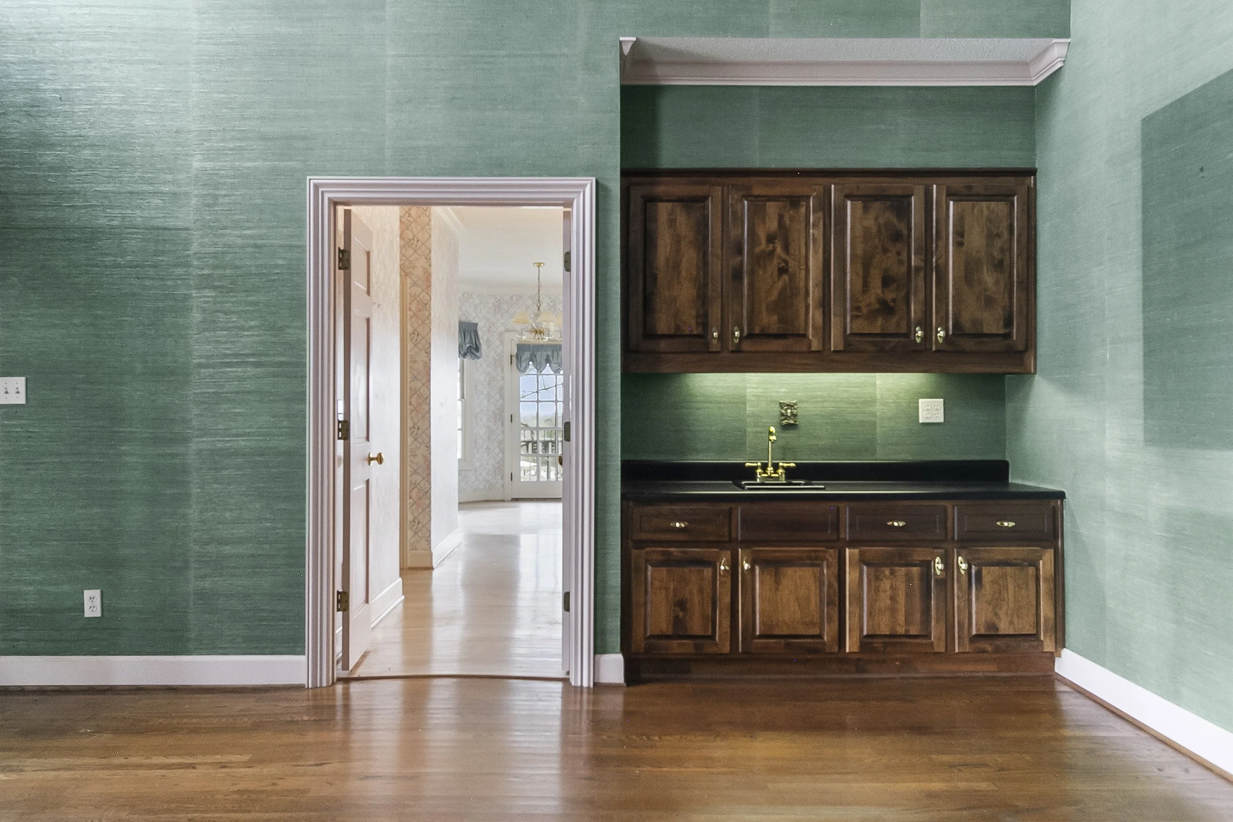 Interior view of a kitchen with green textured walls, wooden cabinets, a black countertop, and a gold faucet. An open doorway leads to a room with patterned wallpaper and a window.