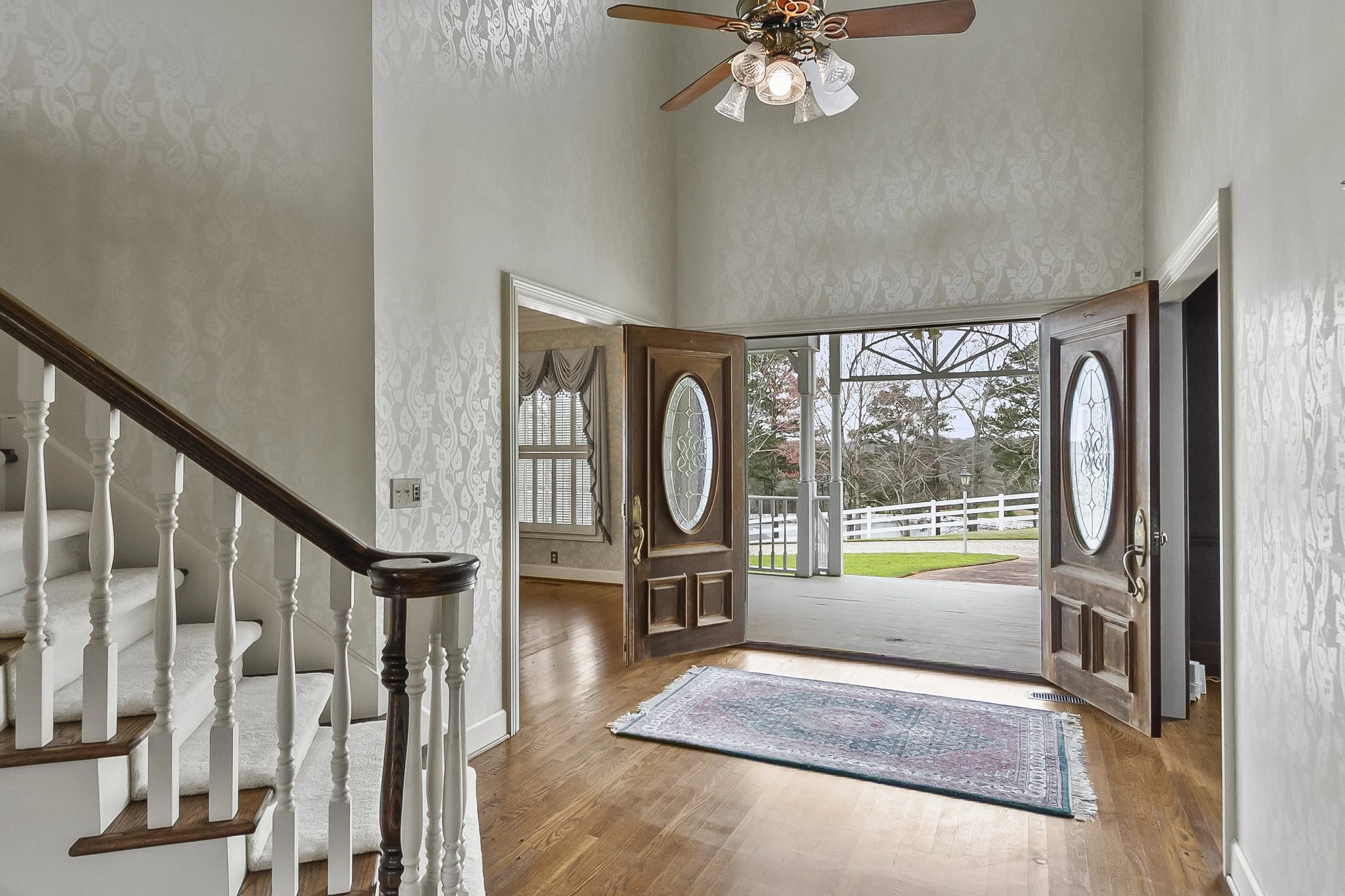 Open front door leading to porch with view of trees and a white fence, interior view of a well-lit entryway with wooden flooring, a rug, a staircase with white and wood banisters, and a ceiling fan with lights.