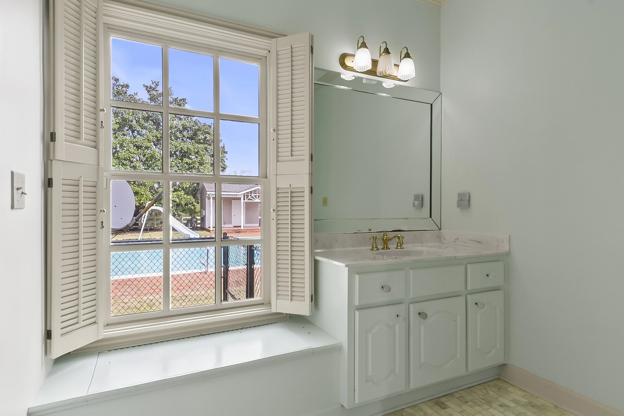 Bathroom with a window, white cabinets, a mirror, and a sink with gold fixtures, overlooking a backyard with a pool and a playground.