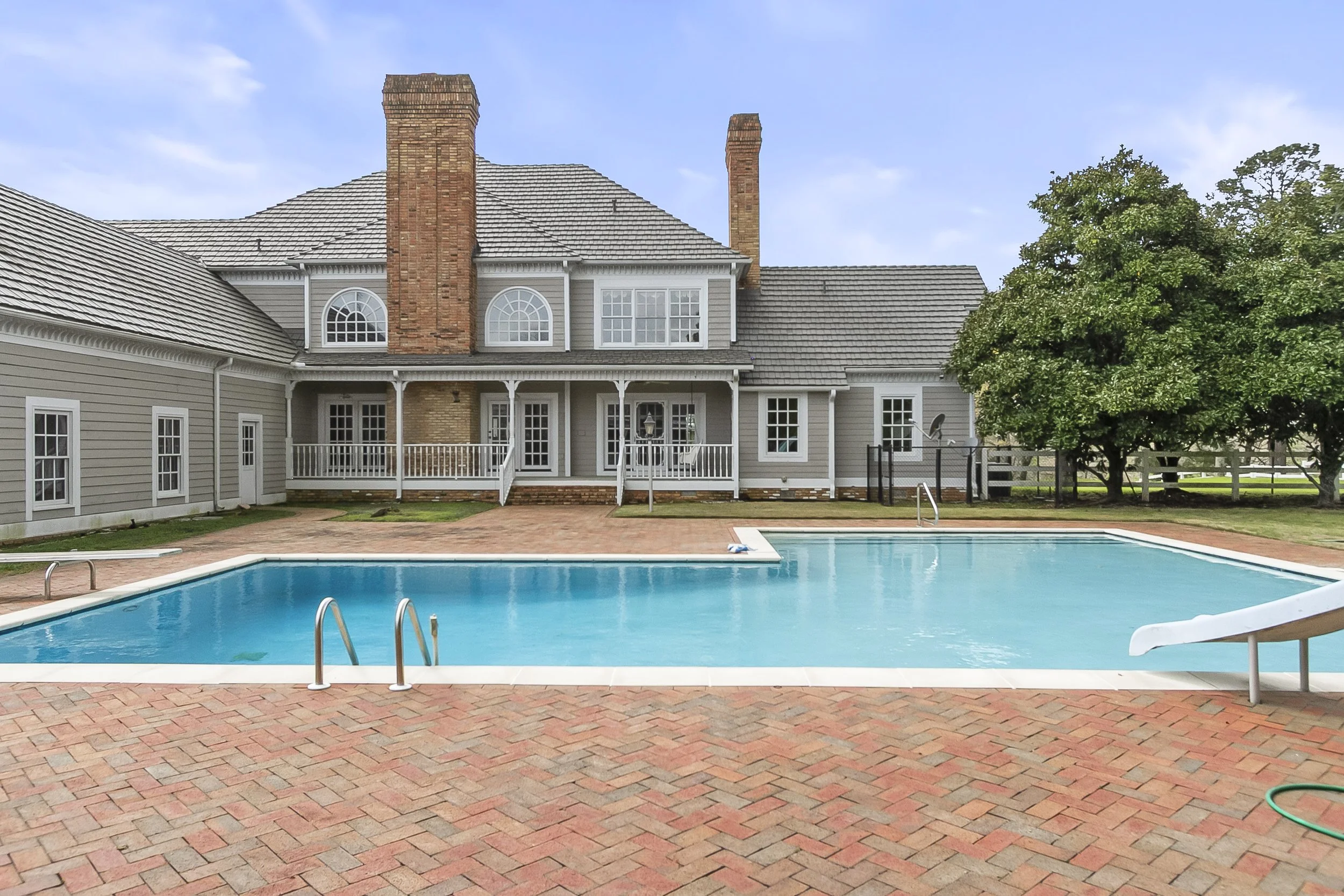 A backyard view of a house with a swimming pool in the foreground, brick patio, and trees on the right side under a partly cloudy sky.