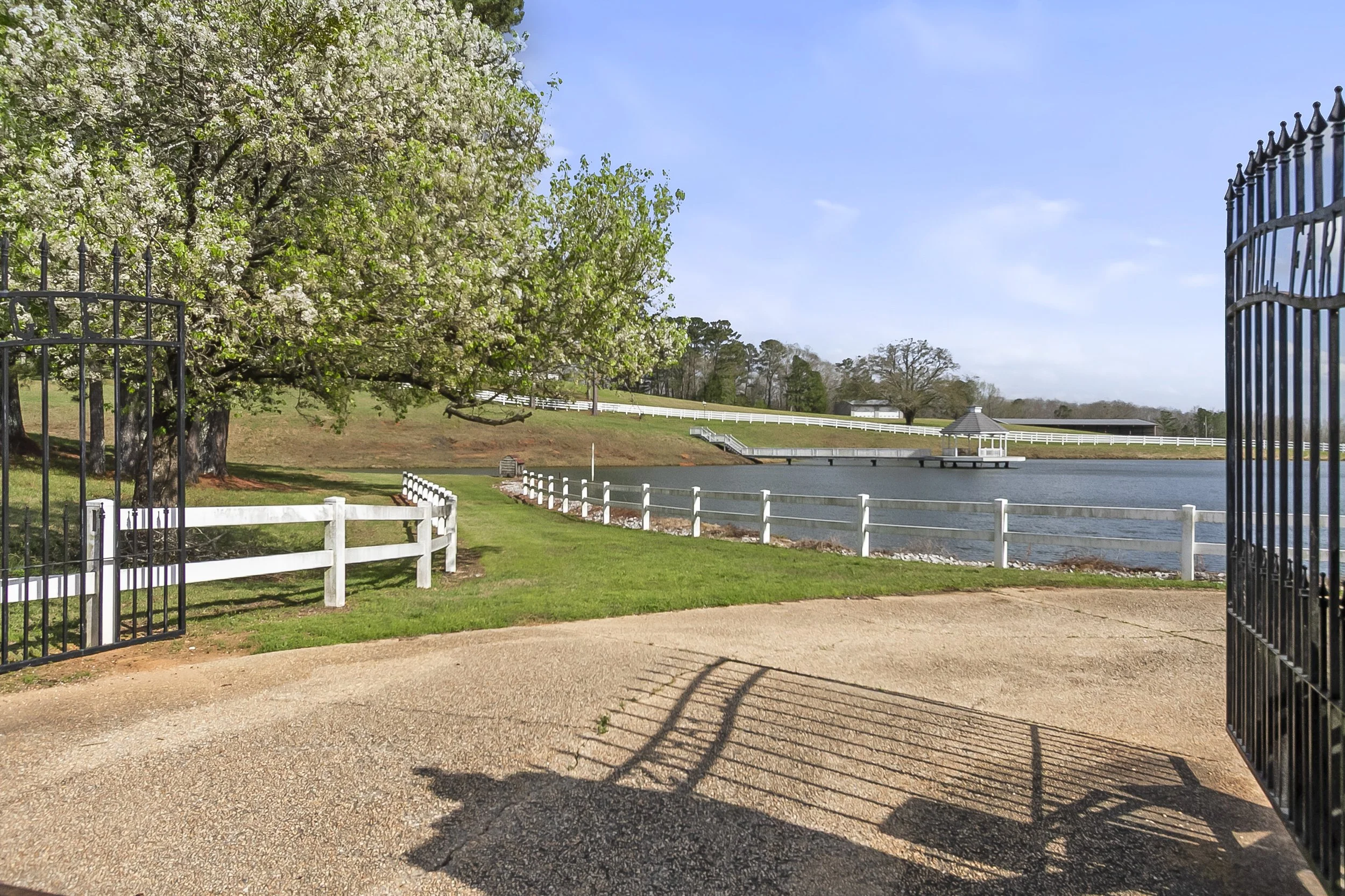A scenic view of a lakeside with a walkway, white fence, and gate in the foreground. A blossoming tree is on the left, and a gazebo is on a small pier over the water. The landscape includes grassy areas, a few trees in the background, and a partly cl