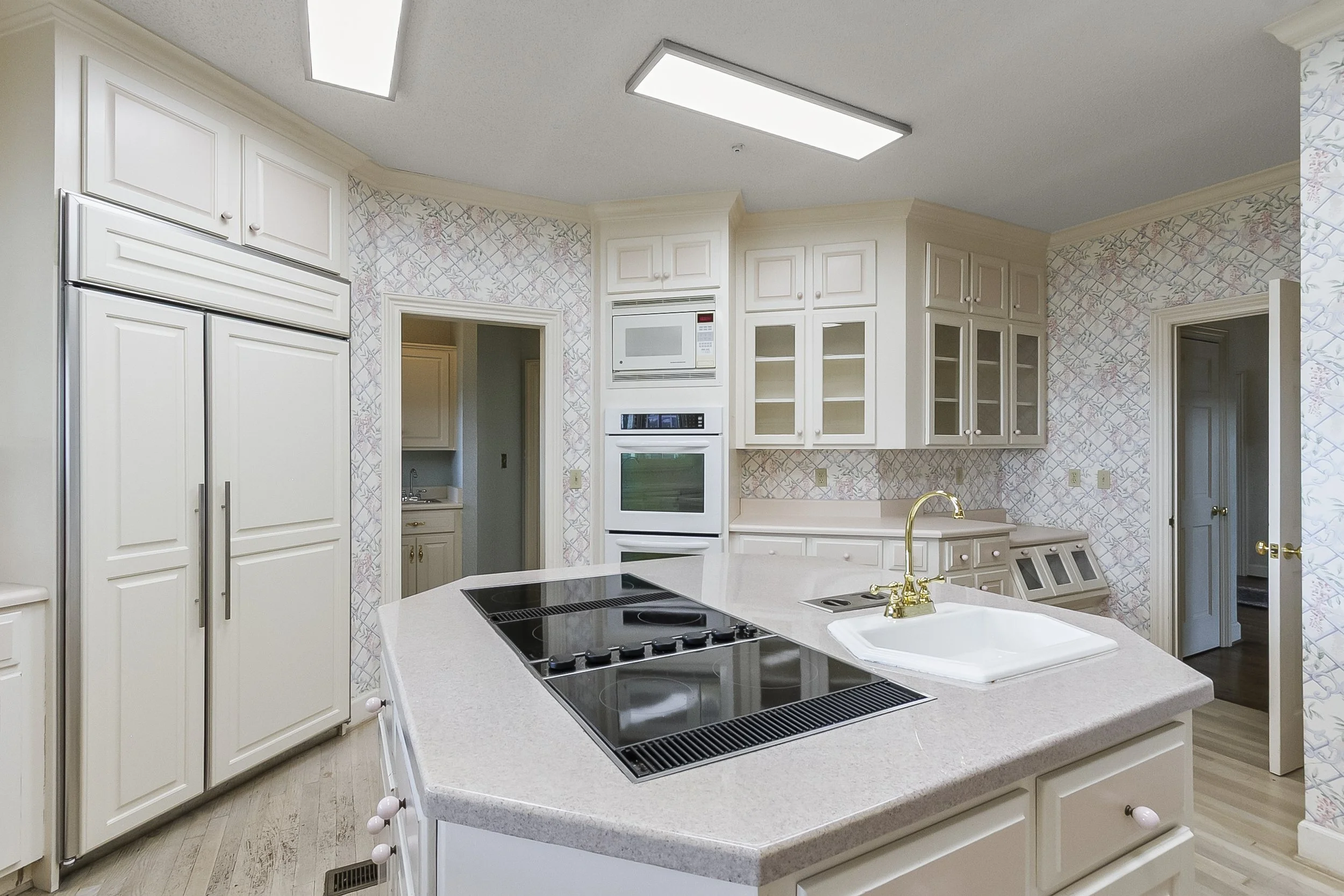 A kitchen with white cabinets, a large island with a built-in stovetop, a gold faucet over a white sink, and a backsplash with a pastel floral and lattice pattern. It has a microwave, double oven, and storage cabinets, with a doorway leading to a sma