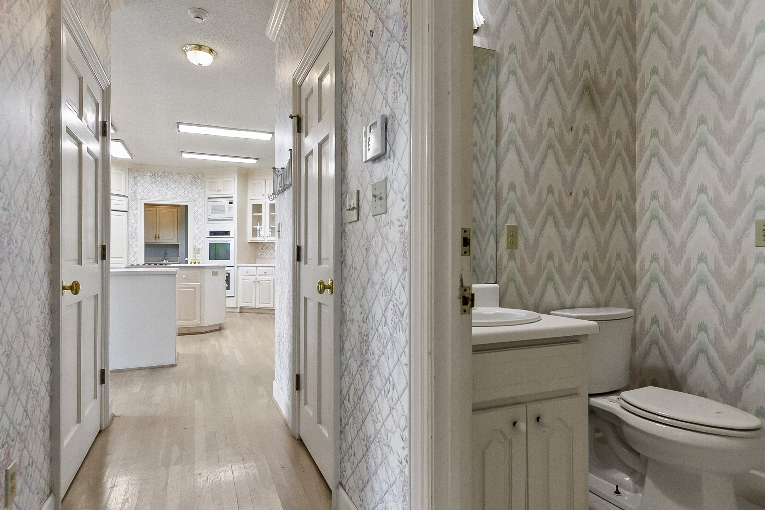 A hallway leading to a white kitchen with cabinets and appliances, and a small bathroom with a toilet, sink, and patterned wallpaper.