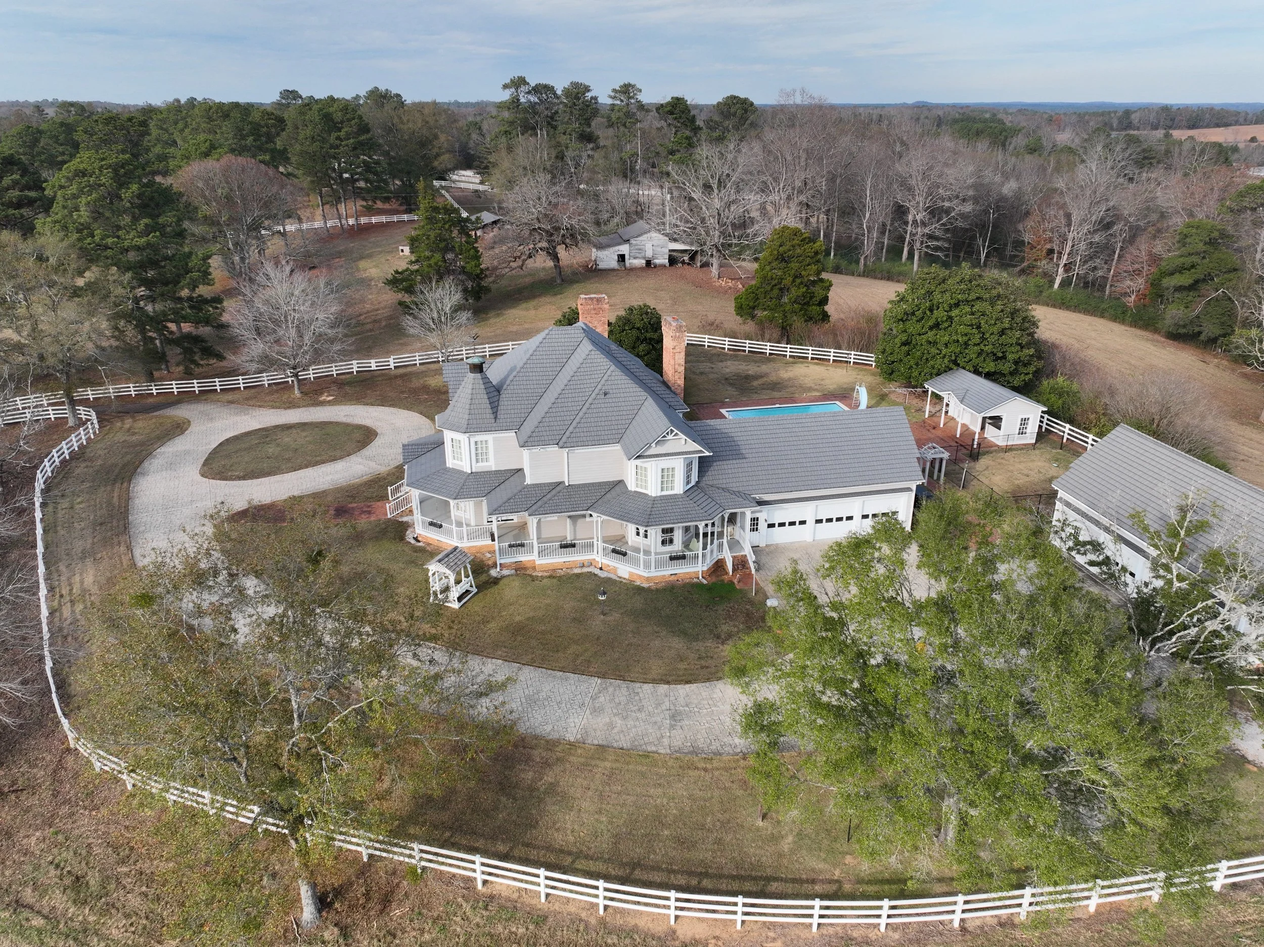 Aerial view of a large property with a main house, a swimming pool, a detached garage, and several outbuildings, all surrounded by white fencing and a mix of trees and open land under a partly cloudy sky.