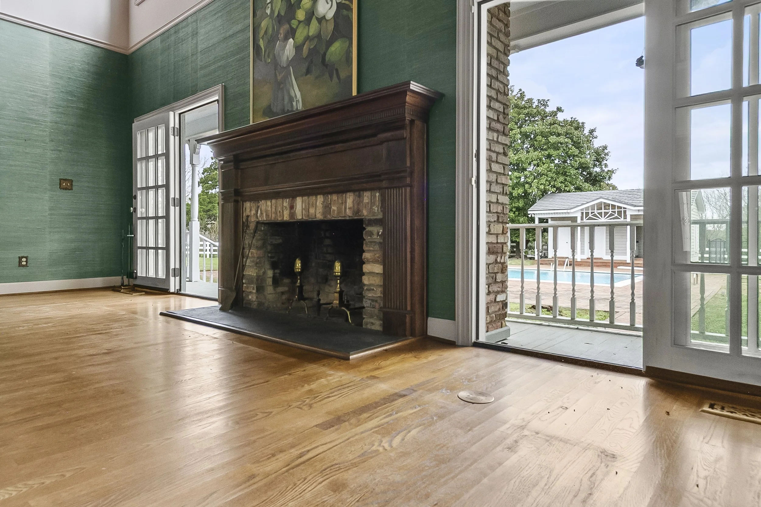 Living room with hardwood floor, green textured walls, a brick and wood fireplace, open French doors leading outside to a porch with a view of a pool and garden area.