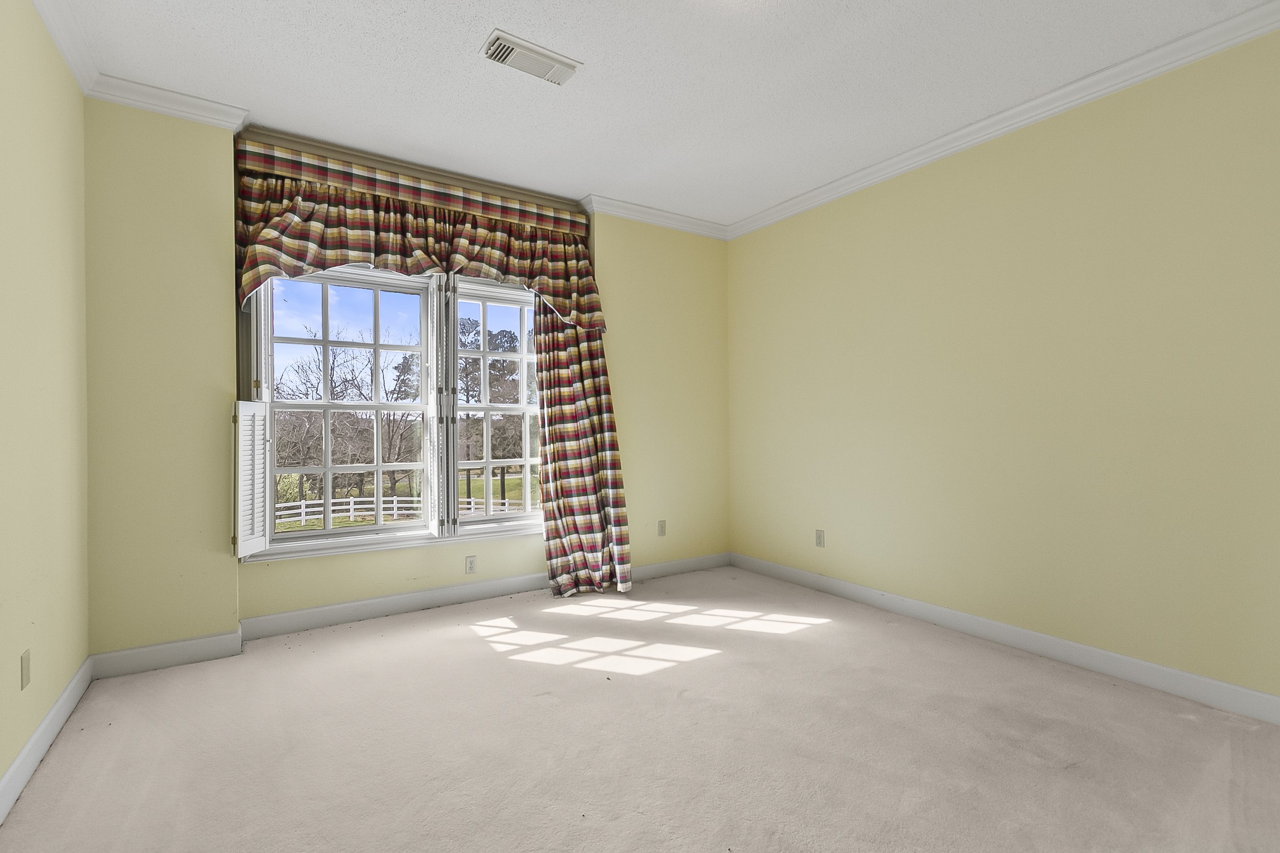 Empty bedroom with yellow walls, white carpet, window with curtains, and sunlight streaming in.