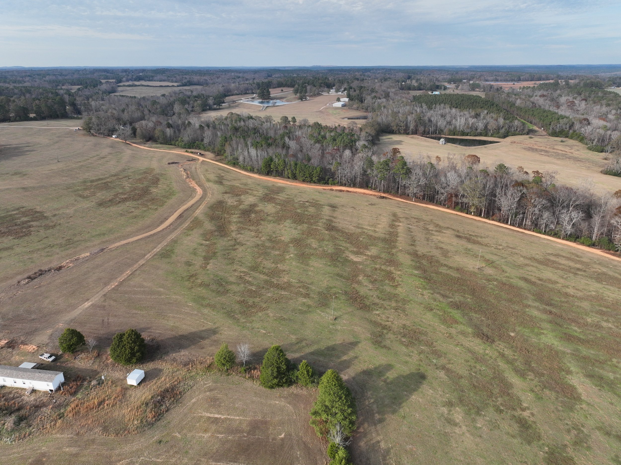 An aerial view of a rural landscape featuring open fields, dirt roads, clusters of trees, and a few scattered buildings, with forested areas in the background under a partly cloudy sky.