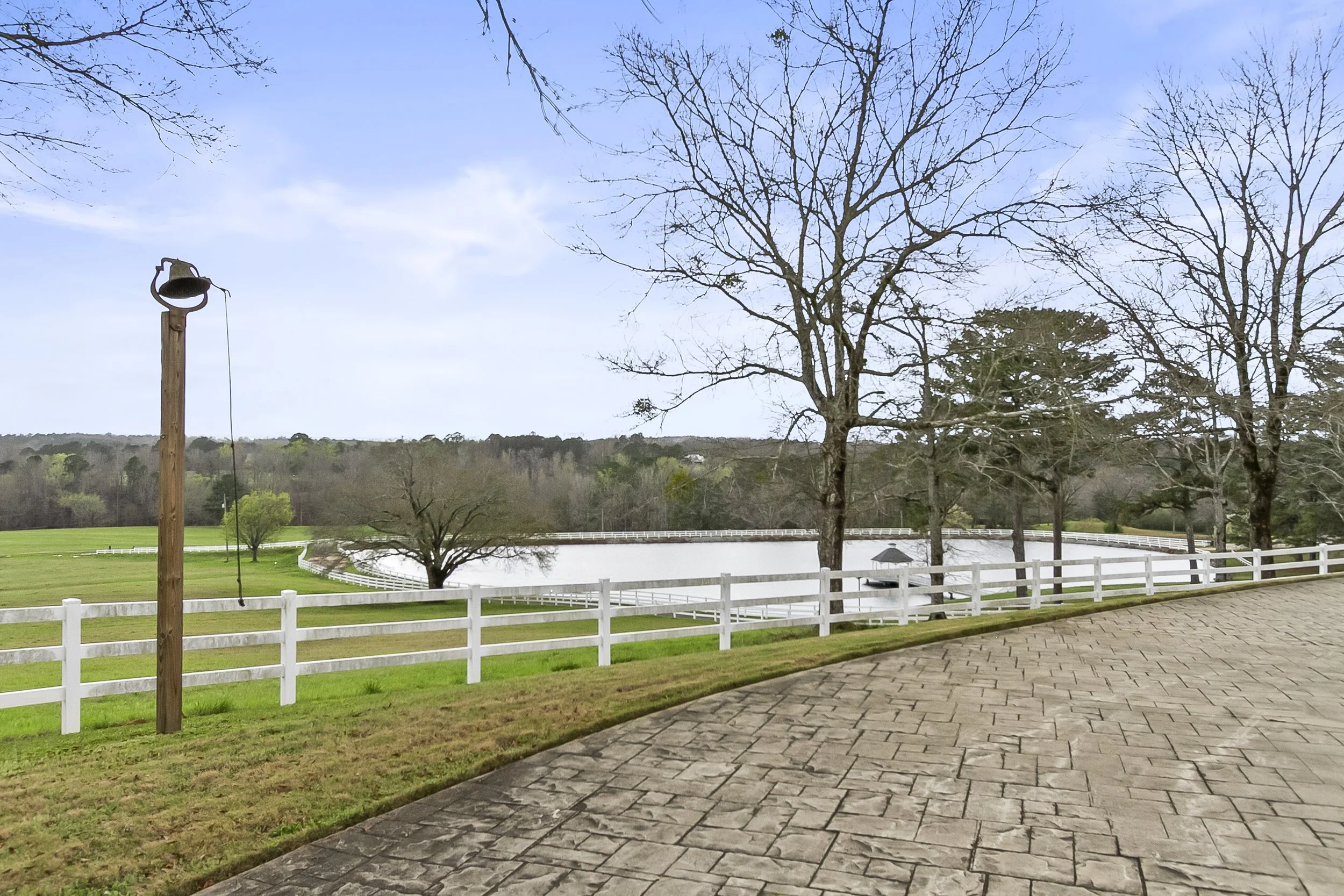 A peaceful outdoor scene featuring a fenced pond in the distance, leafless trees, and a wrought iron gazebo under a partly cloudy sky, with a stone paved pathway in the foreground.