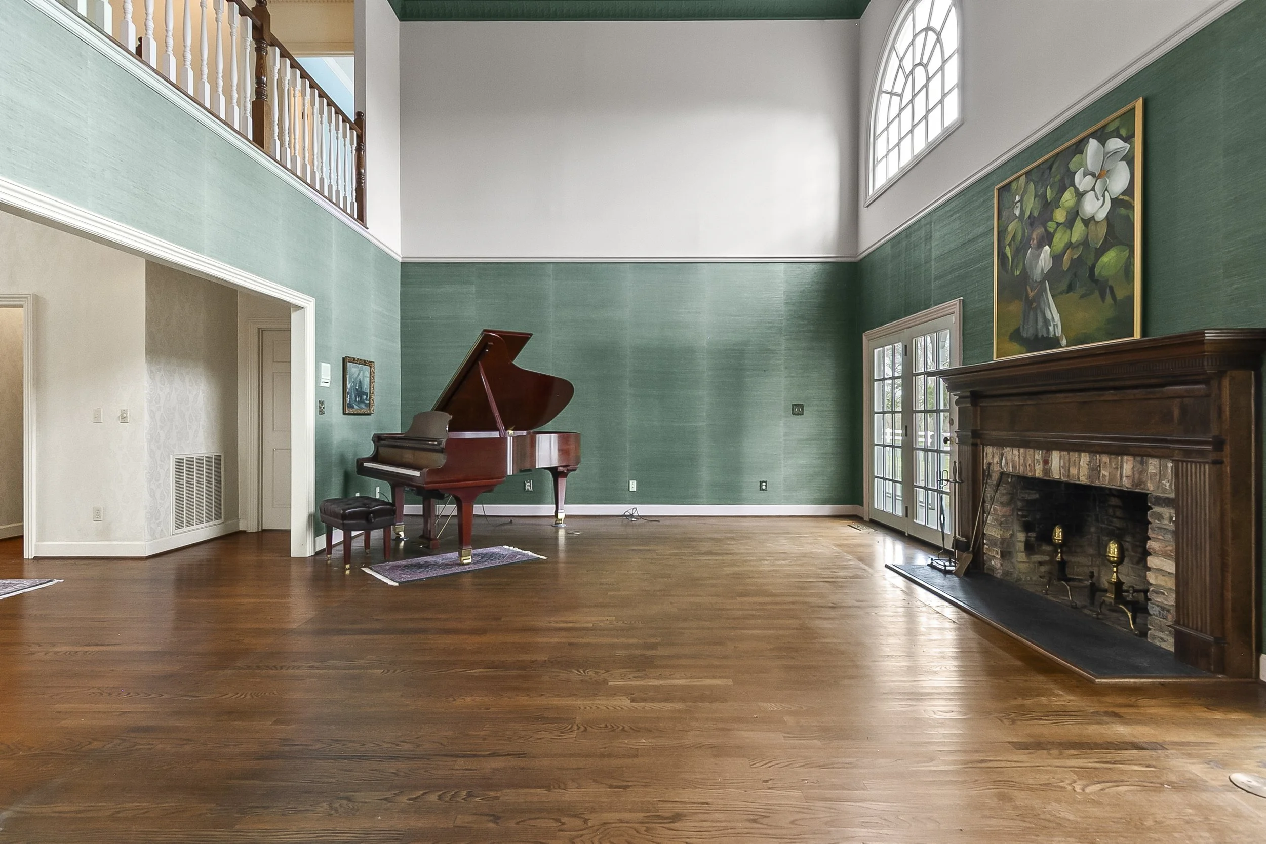 Spacious living room with a grand piano on the left, wooden floors, green textured walls, a large picture window with an arched top on the right, and a brick fireplace with a wooden mantel on the far right.
