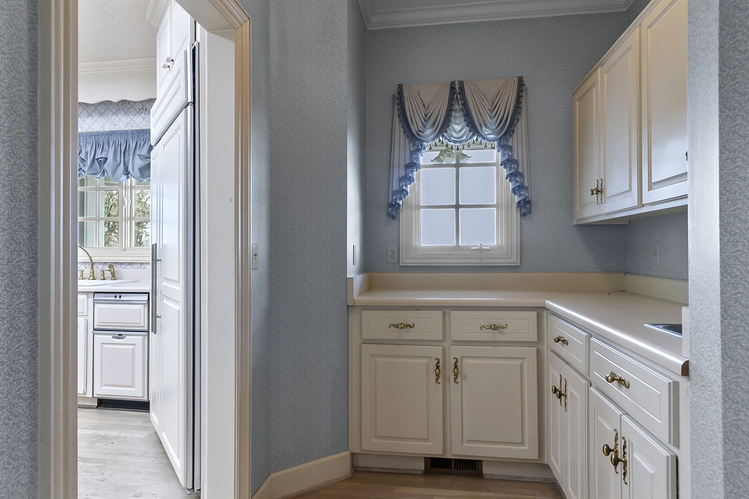 Kitchen with window, white cabinets with gold hardware, blue and white curtains, and beige countertops.